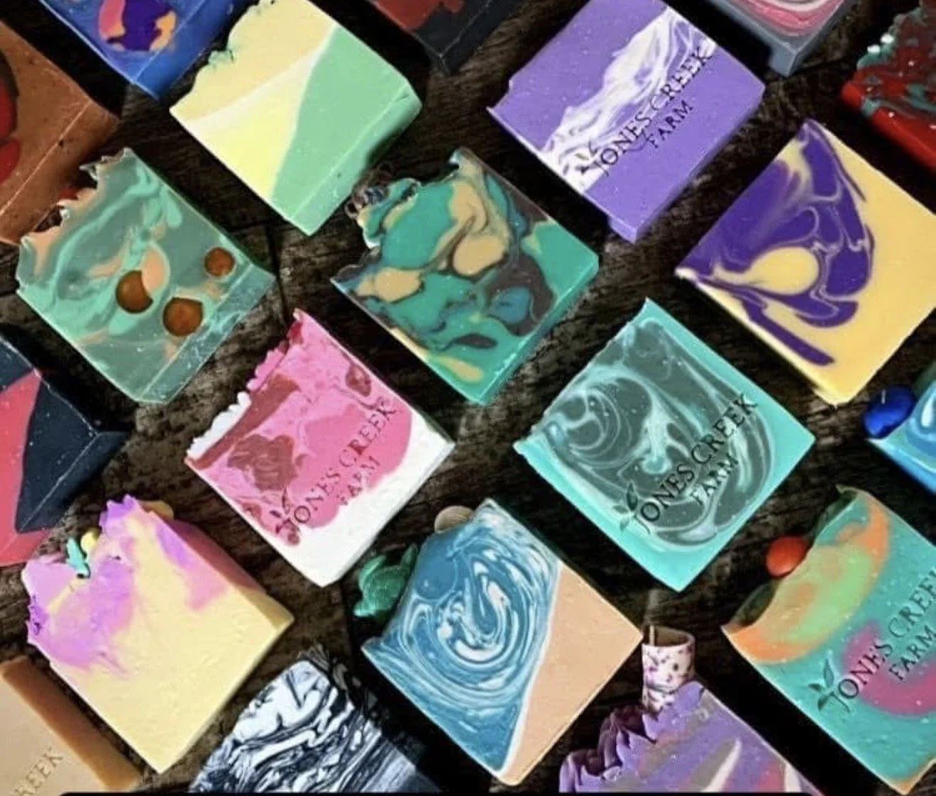 Colorful handmade soap bars with marbled and swirl patterns displayed on a wooden surface, some with the 'Joan's Creek Farm' logo.