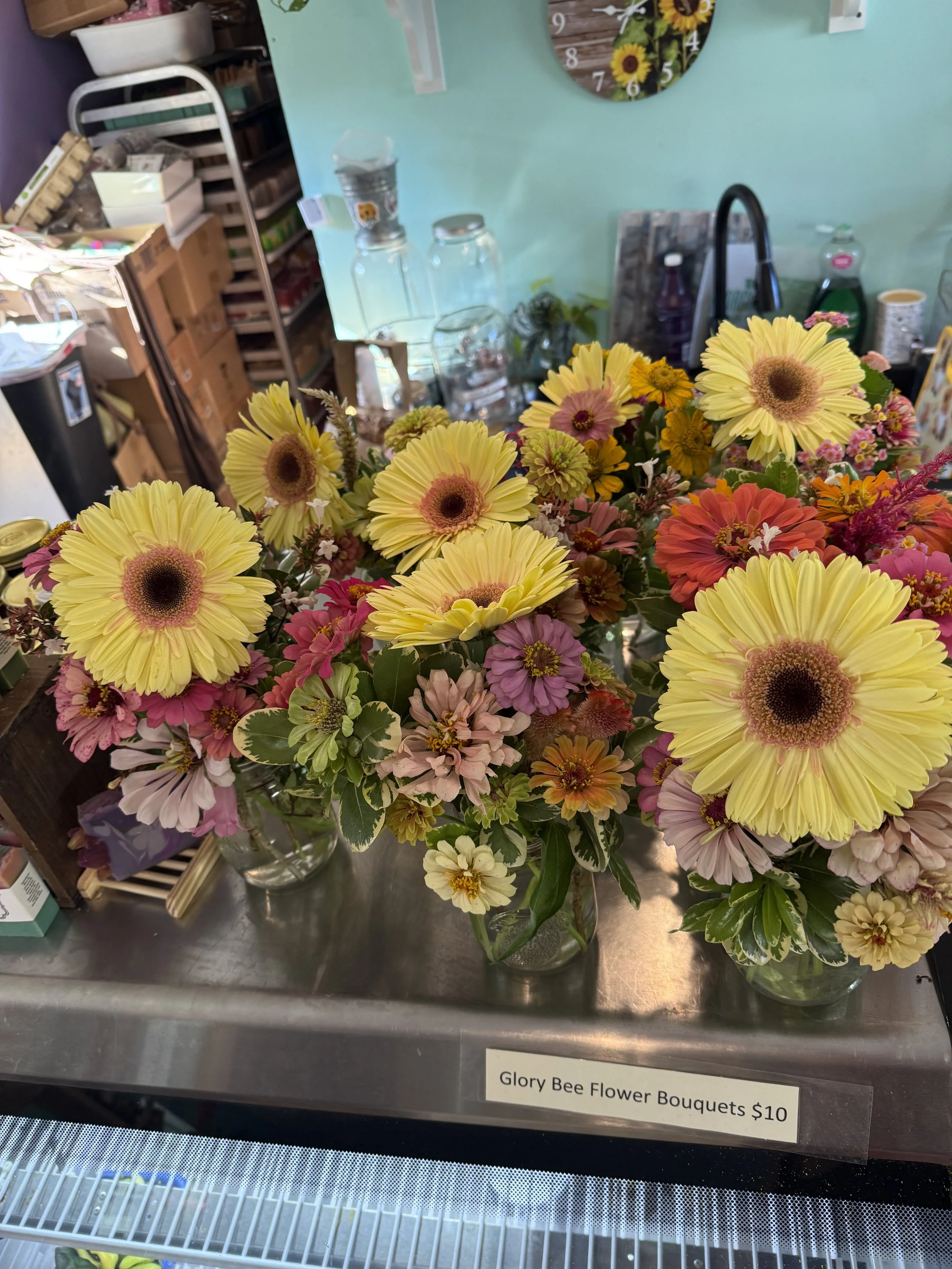 A bouquet of yellow, pink, and orange flowers in a clear glass vase on a metal surface, with a label reading 'Glory Bee Flower Bouquets $10', in a store with shelves holding various items in the background.