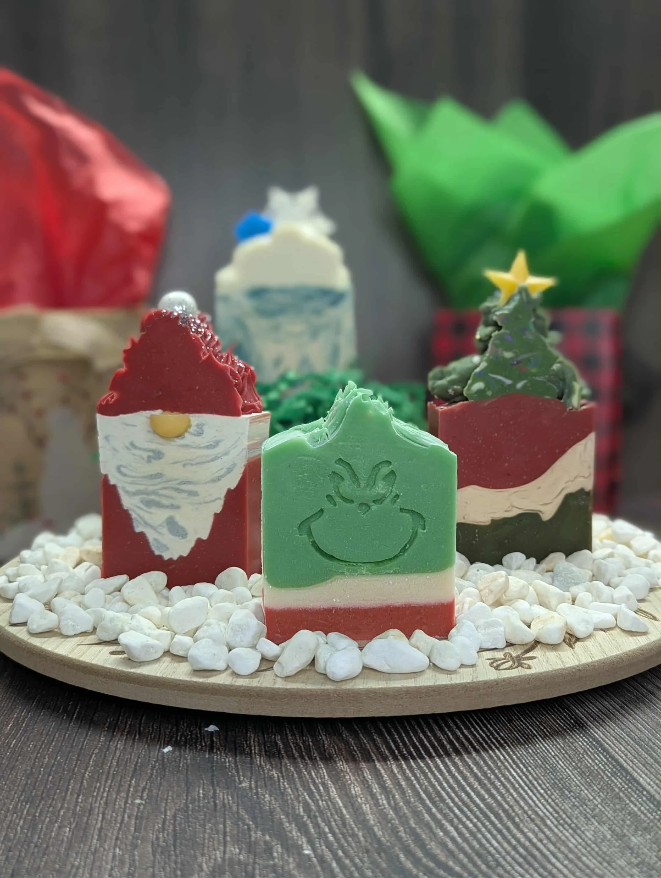 Decorative soaps with Christmas themes arranged on a round wooden tray filled with white pebbles, set on a dark surface.