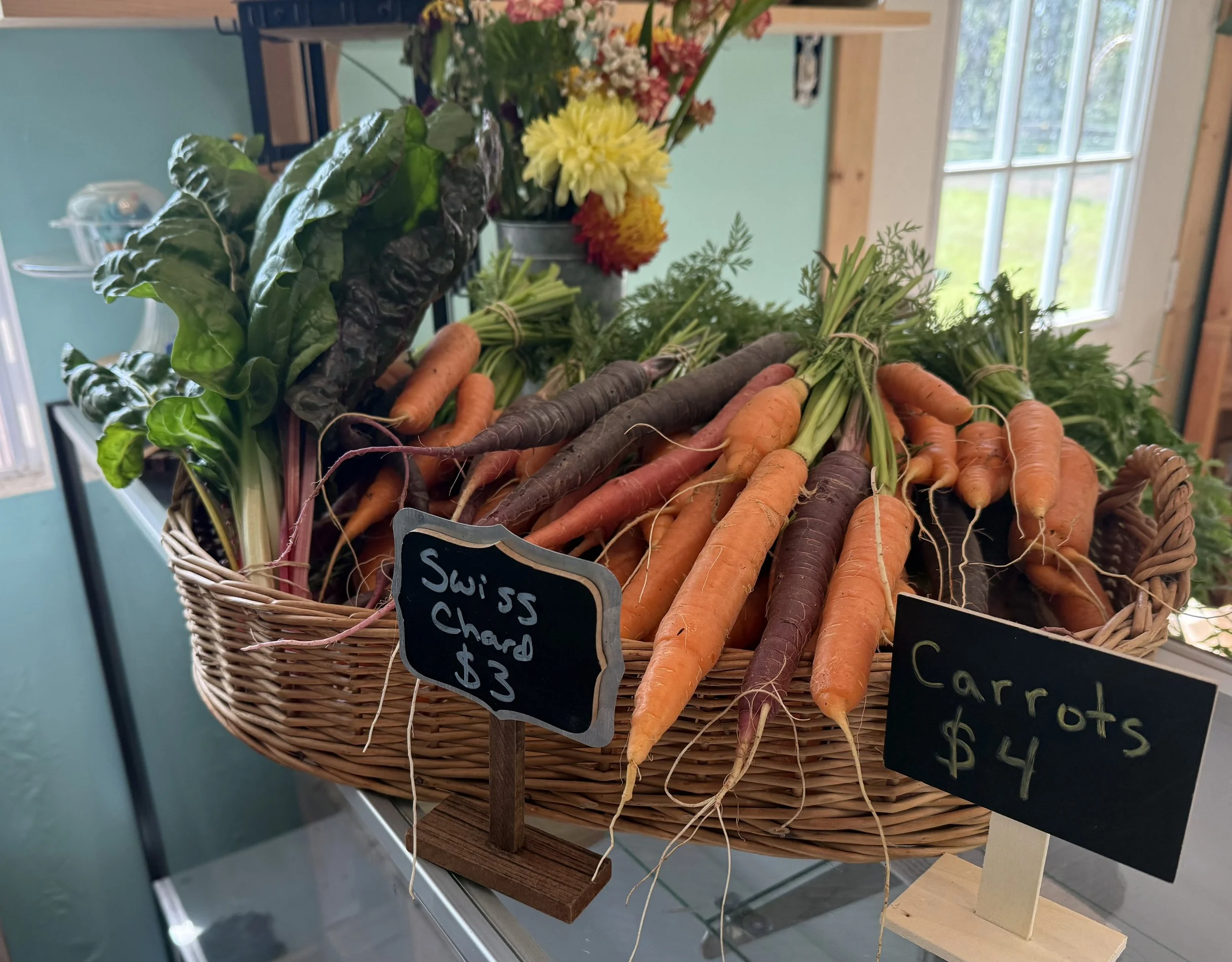 A basket of fresh carrots and leafy greens at a farmers market stall, with small handwritten signs displaying prices.
