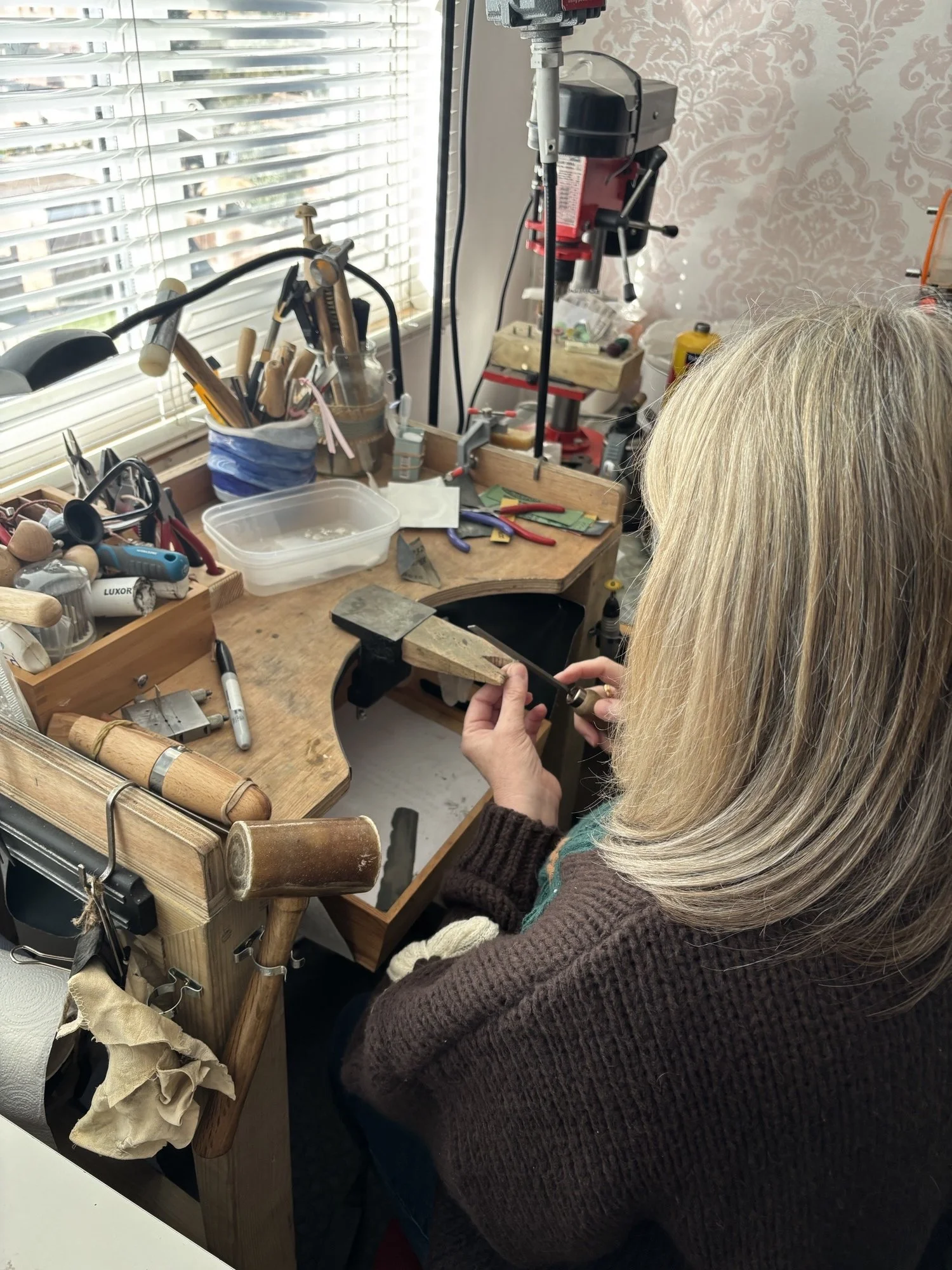 Close-up of a person working at a woodworking workbench with various tools, a drill press, and wood pieces, in a workshop with natural light coming through window blinds.