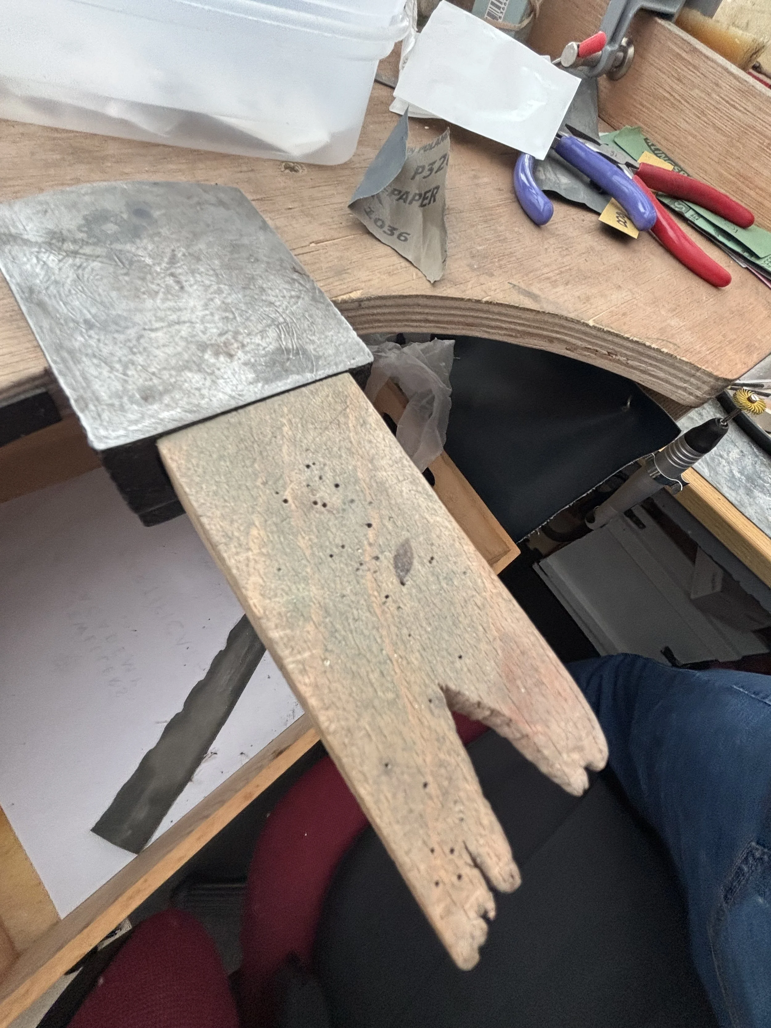 A wooden workbench with various tools including pliers, a metal clamp, sandpaper, and colored papers, in a workshop setting.
