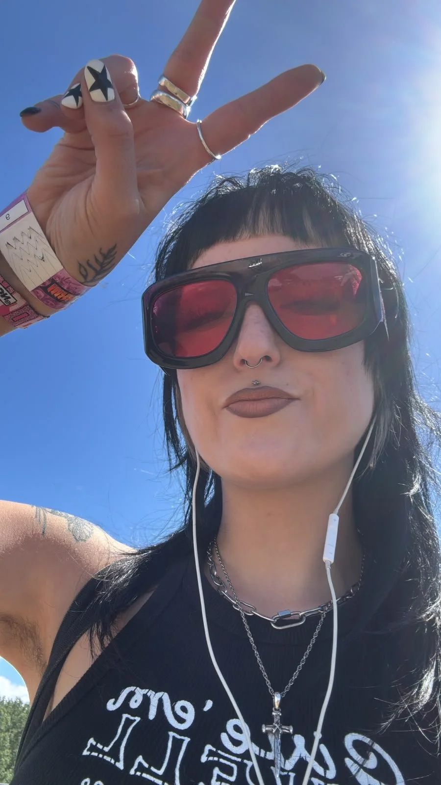 A person outdoors wearing sunglasses and earbuds, making a peace sign with their hand, dressed in a black tank top and accessories, against a bright blue sky.