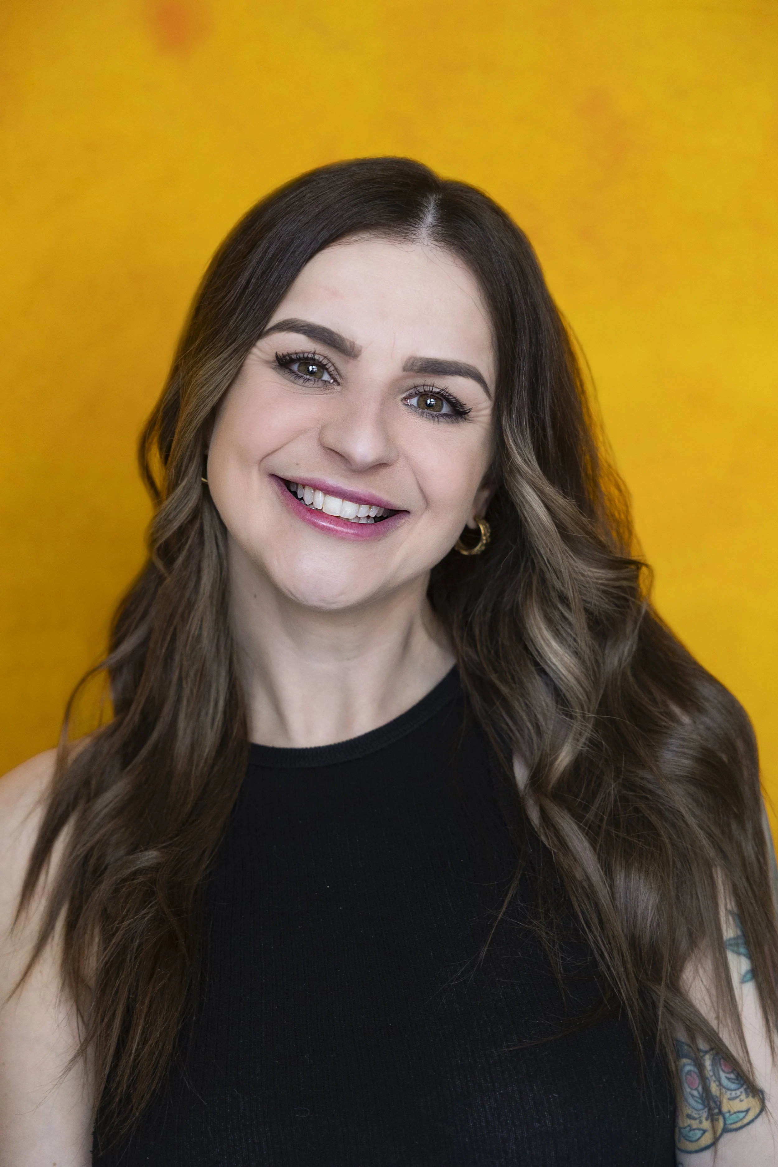 A woman with long, wavy brown hair and a bright smile, wearing a black sleeveless top, standing against a bright yellow background.