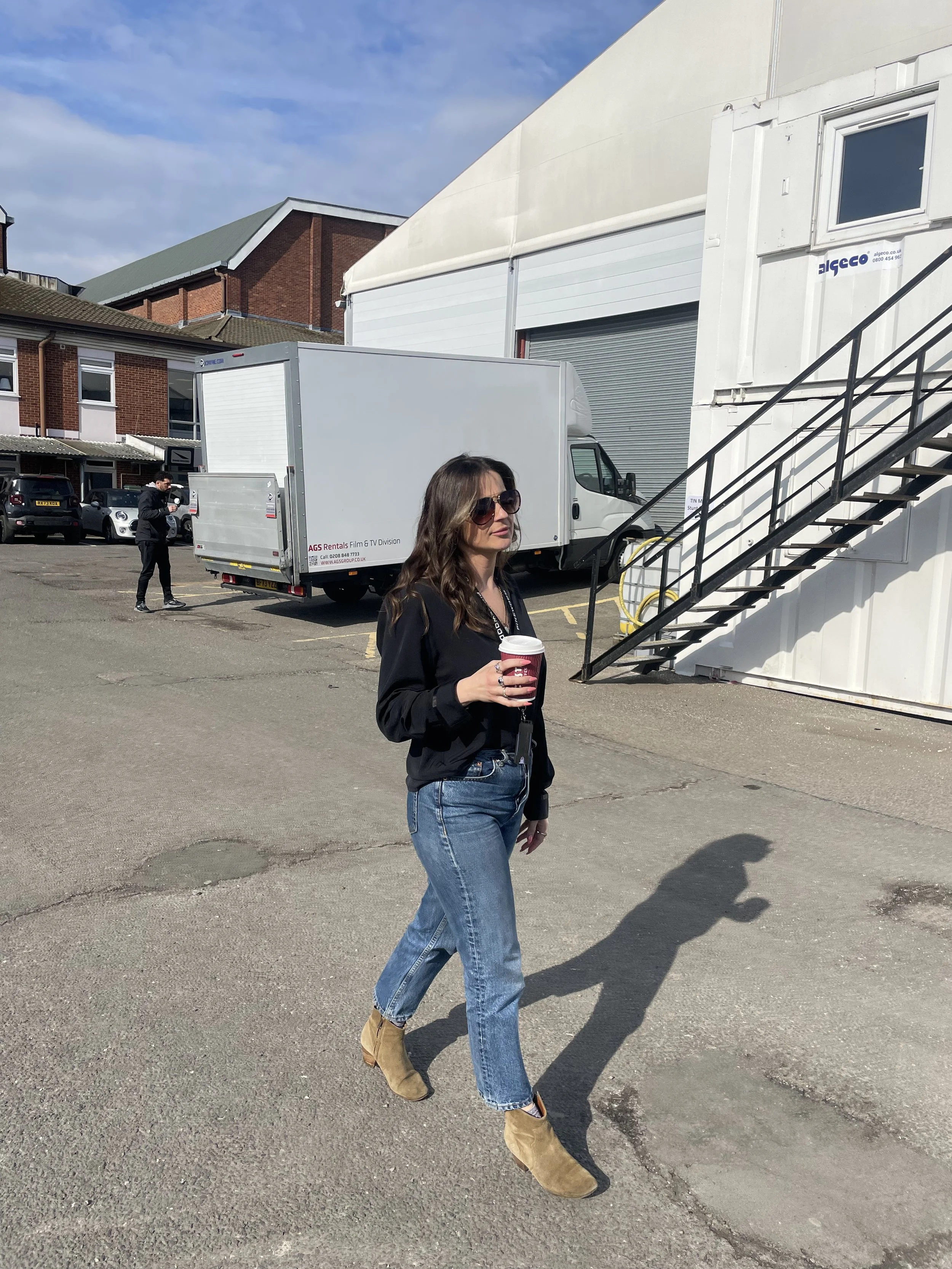 A woman with long brown hair wearing sunglasses, a black jacket, blue jeans, and brown boots holding a coffee cup walking outdoors in a parking lot.