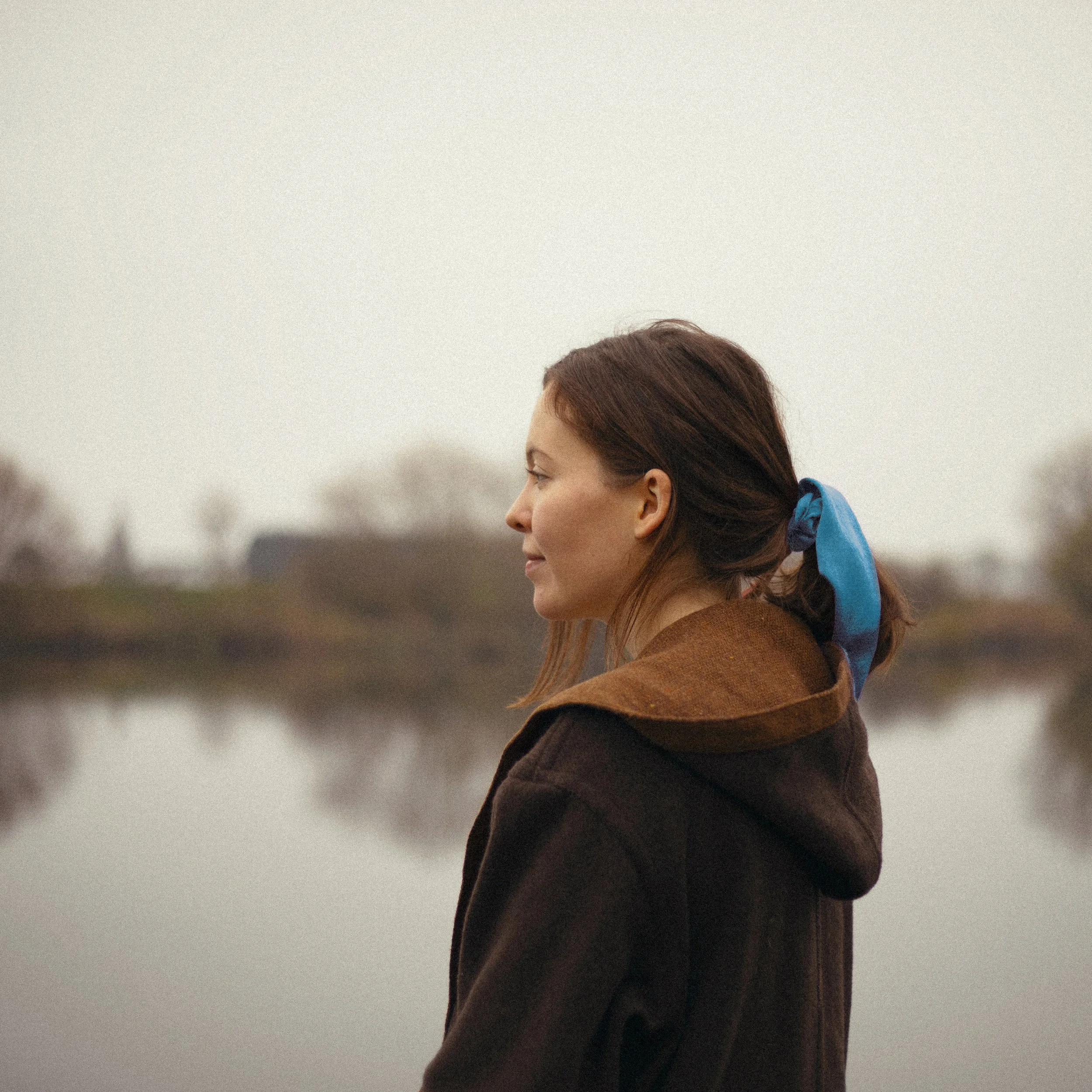 A profile view of Amy Fitzgerald with brown hair tied back with a blue hair tie, wearing a brown coat, standing outdoors with a blurry background of trees, lake and cloudy sky.