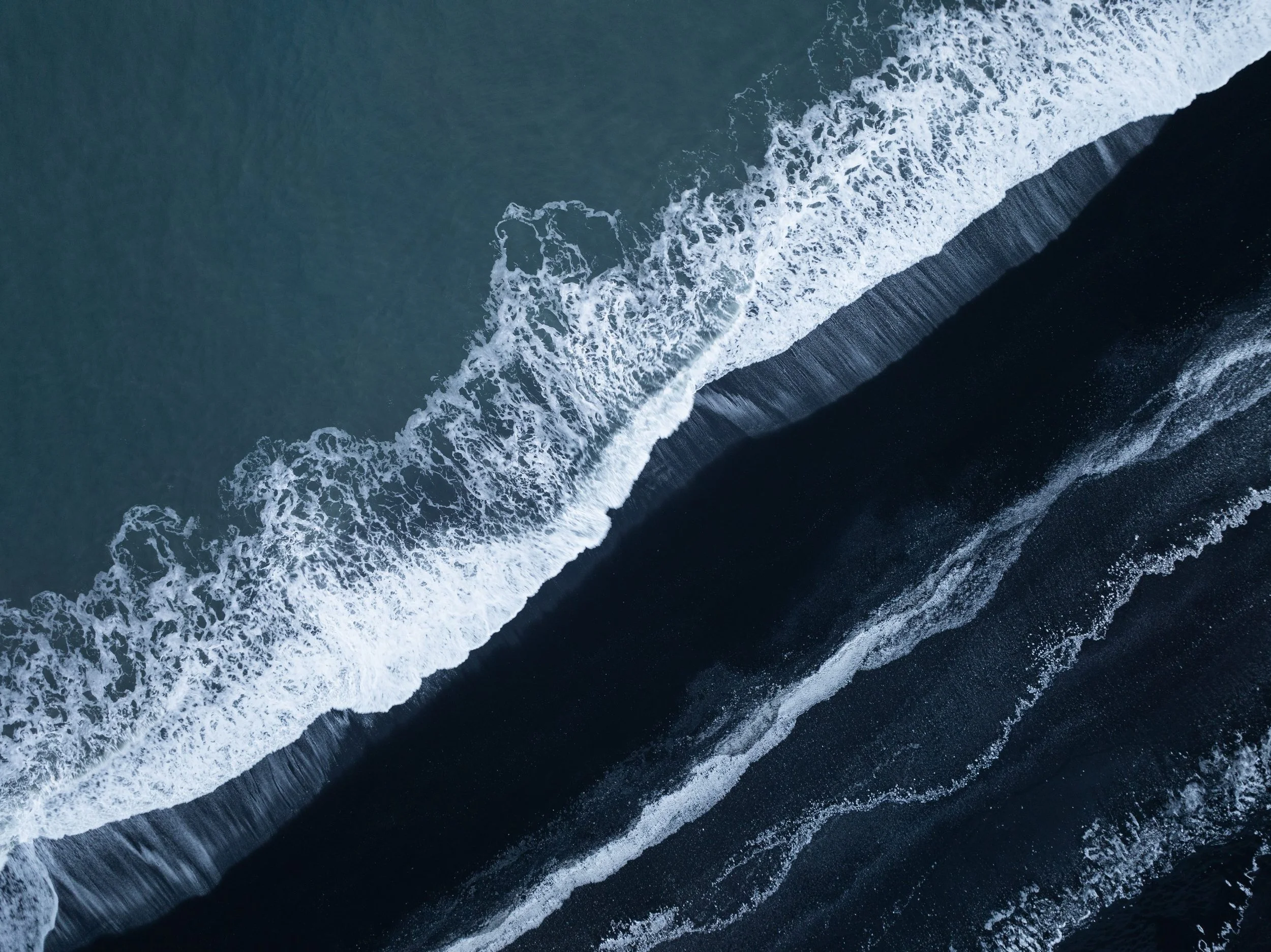 Aerial view of ocean waves meeting a black sand beach, a line dividing sea from shore.