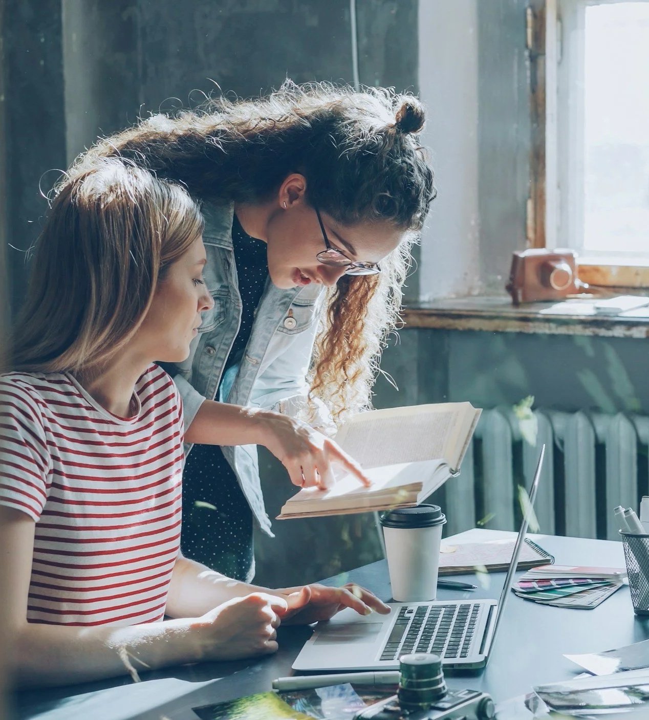 Twee vrouwen die samen naar een geopend boek kijken aan een houten tafel.