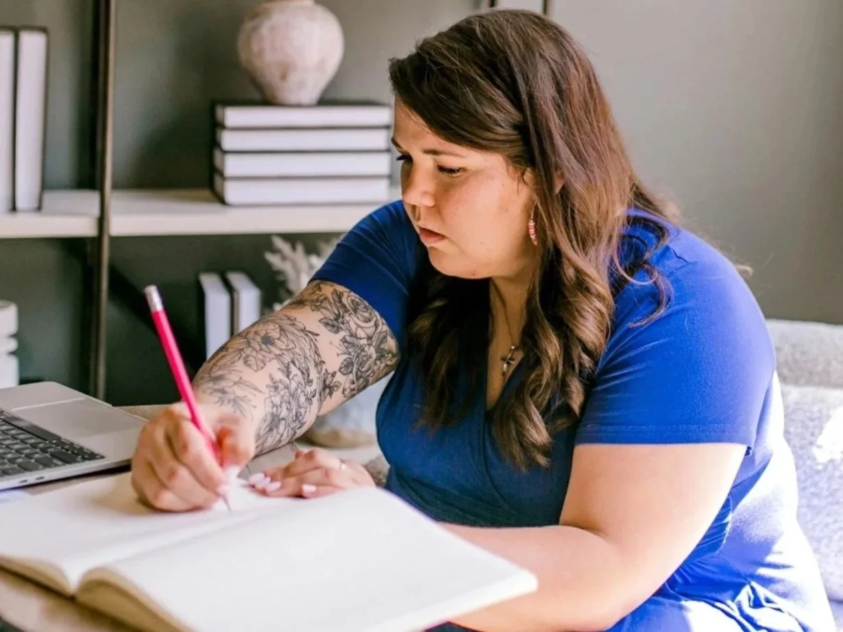Een vrouw schrijft met de hand in een notitieboek, met een laptop en telefoon op tafel.