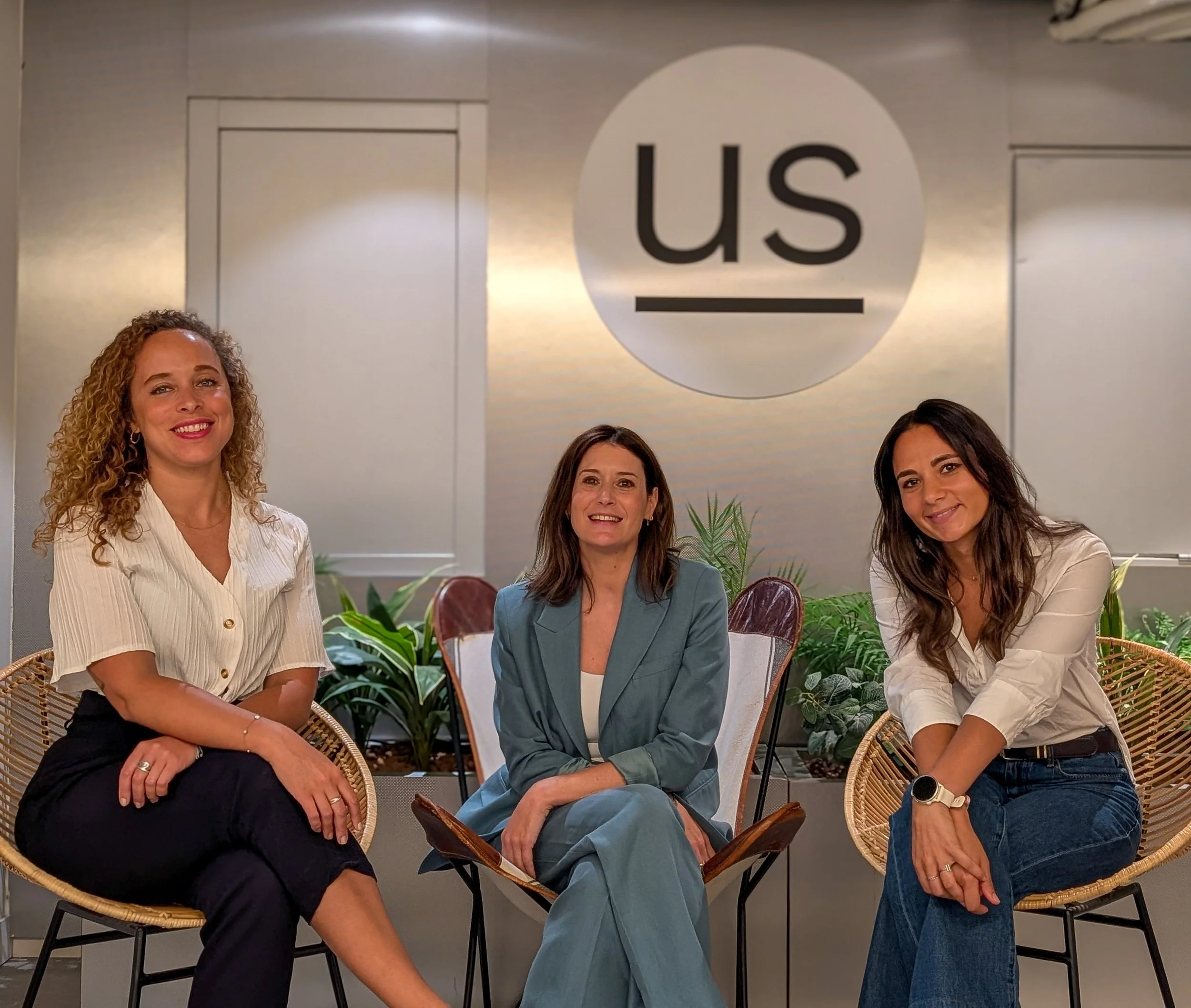 Quatre femmes assises en groupe dans une salle d'attente ou une réunion, avec un panneau indiquant 'US' derrière elles, entourées de plantes vertes.
