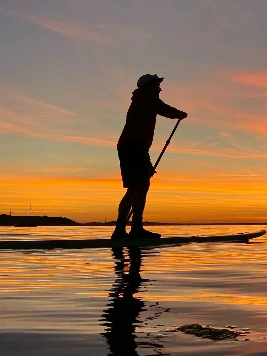 Silhouette of Jeremy Clark paddleboarding in Poole Harbour during a colorful sunset, with reflections on the water and a distant shoreline.