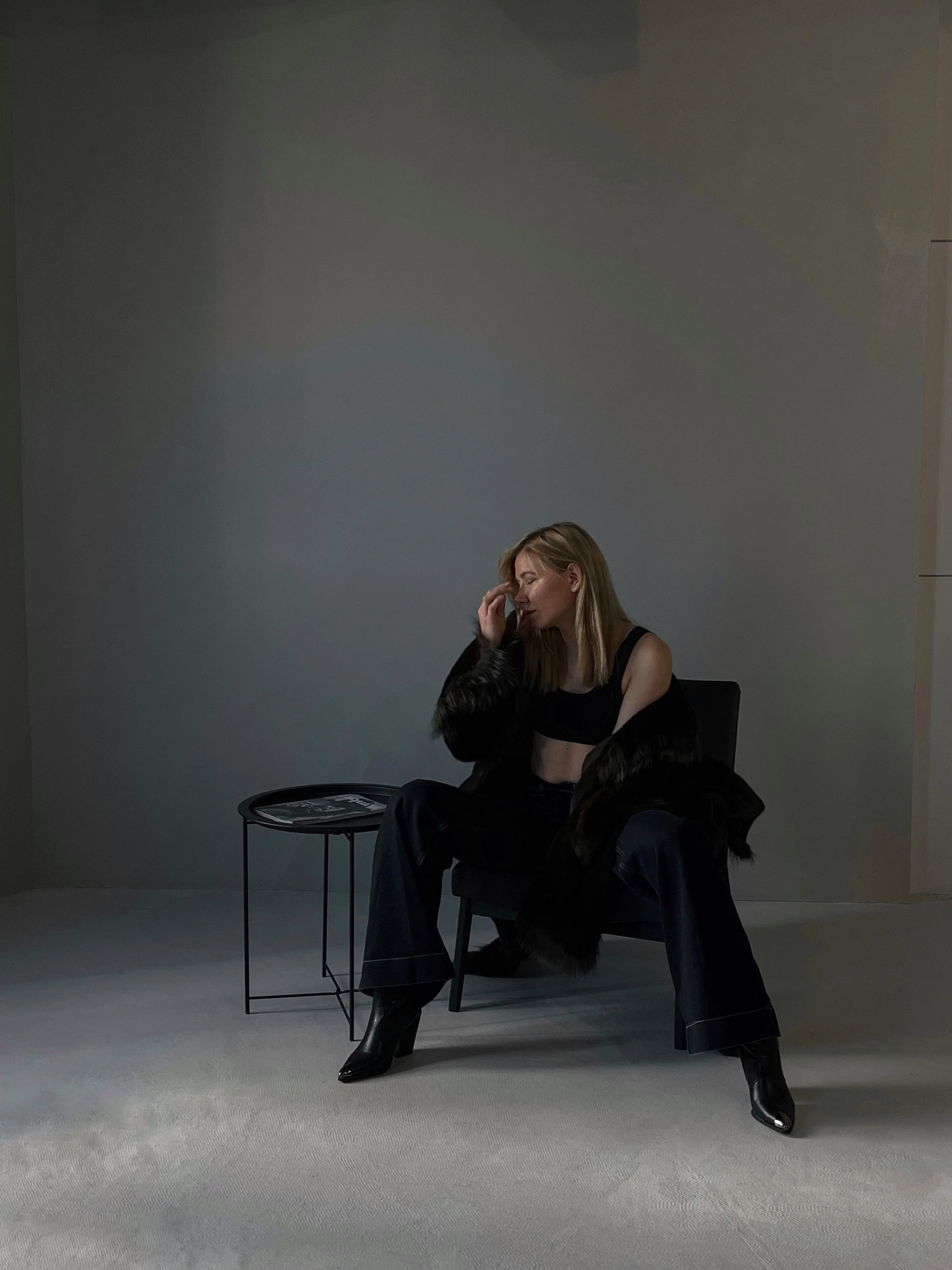 A woman with blonde hair, wearing a black crop top, black pants, and shiny black boots, sitting on a black chair with one leg crossed over the other, with a second person sitting on her lap. She is touching her forehead with her right hand, and the background features a plain wall, a small black side table, and a book on top.
