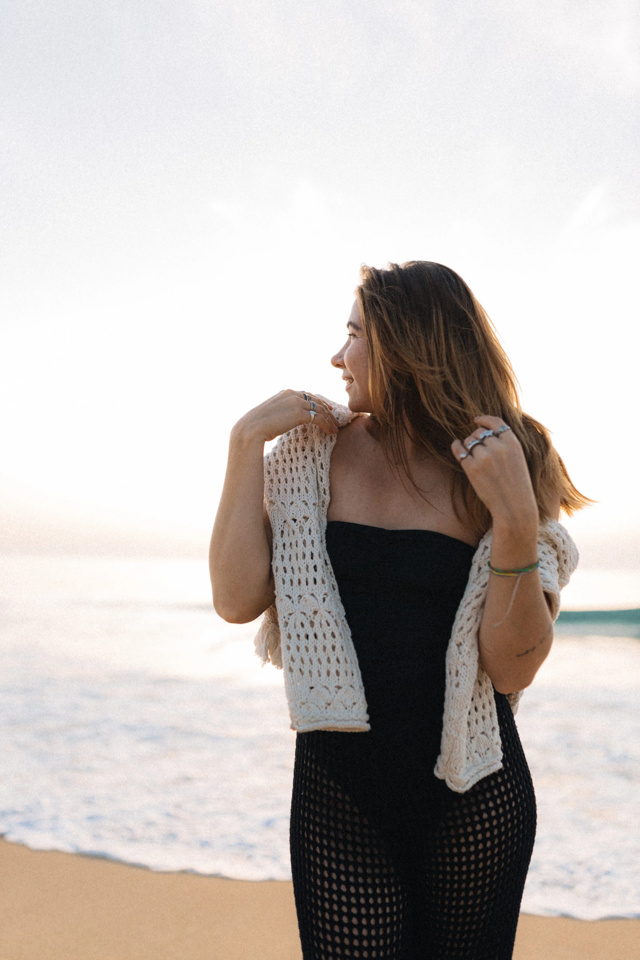 A woman with long, reddish-brown hair standing on the beach, smiling, with her eyes closed, wearing a black dress and a beige crochet cover-up, during sunset.