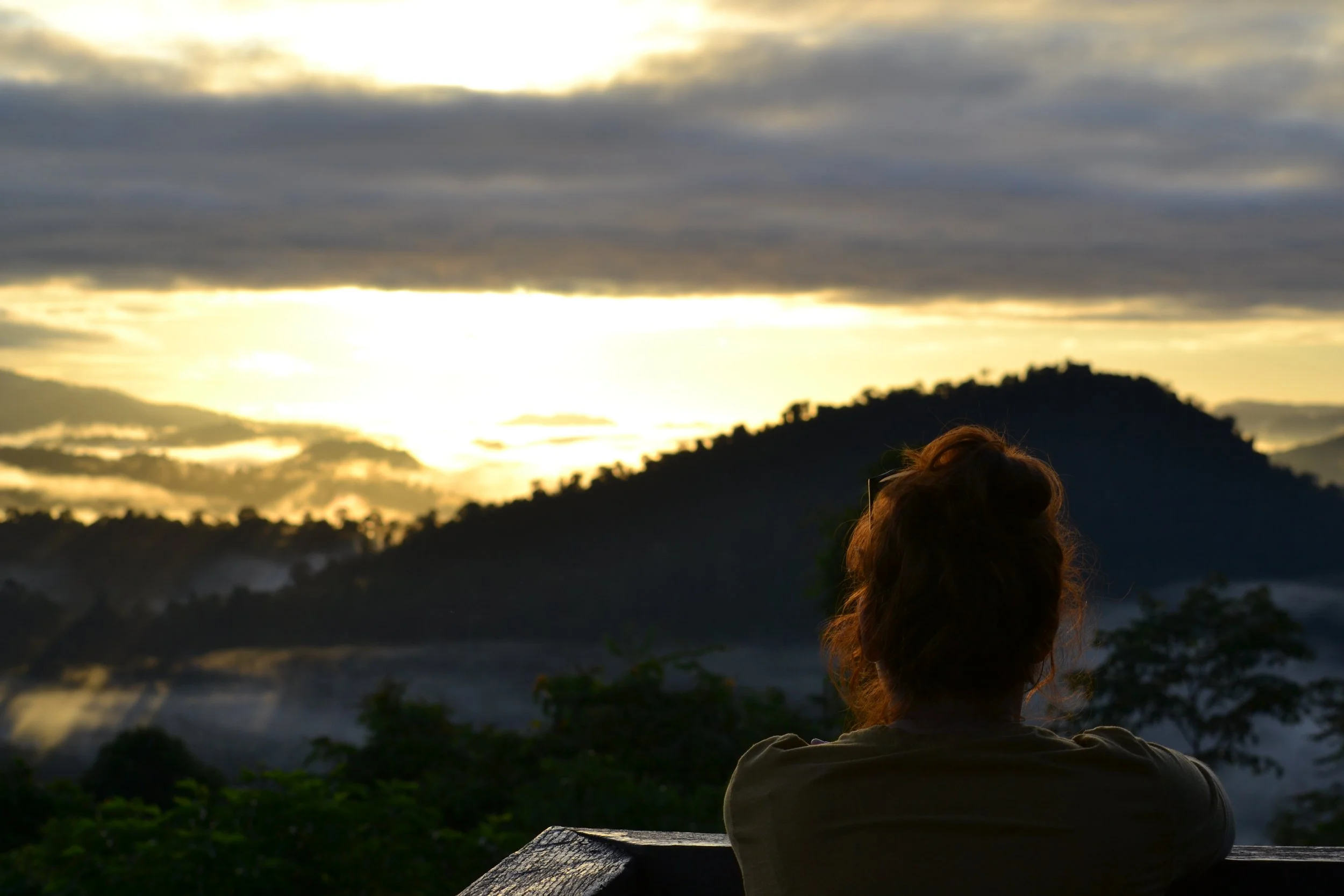 A person with reddish hair sitting on a wooden bench, gazing at a sunset with a mountainous landscape and cloudy sky in the background.