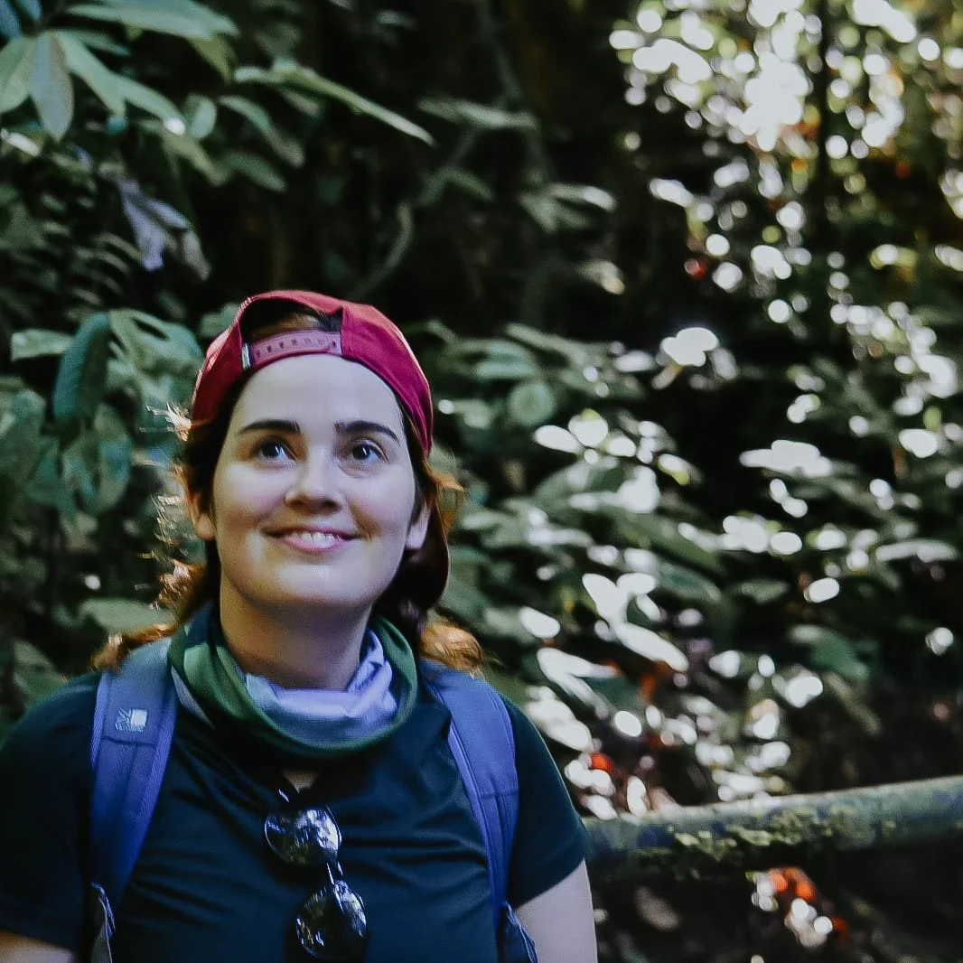 Young woman with red hair, wearing a red cap backwards, a black t-shirt, and a green neck gaiter, smiling in a lush forest with dense green foliage.