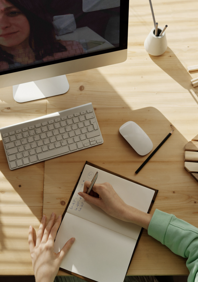 Bureau en bois avec un ordinateur, un clavier, une souris, un carnet avec une personne écrivant, un stylo et des objets de bureau en blanc.