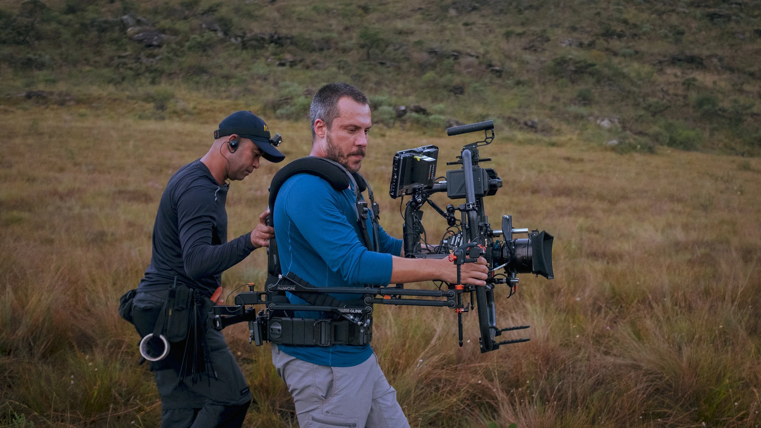 Two men operating a professional camera mounted on a stabilizer rig outdoors in a grassy field with a hillside in the background.