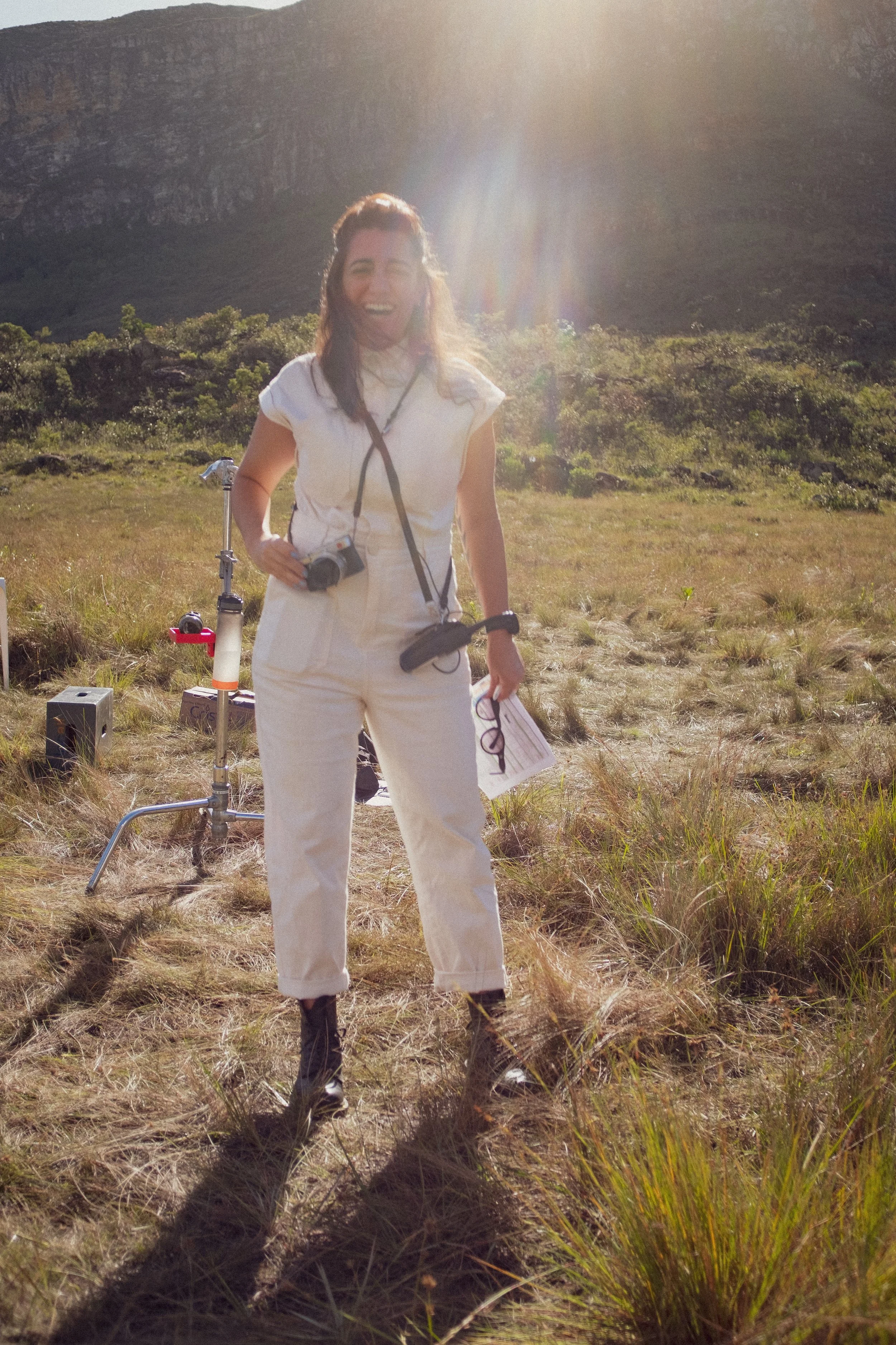 A woman standing outdoors in a grassy field with mountains in the background, smiling, with a camera hanging around her neck, and holding a piece of paper or notebook in her left hand. The sun is shining brightly behind her.