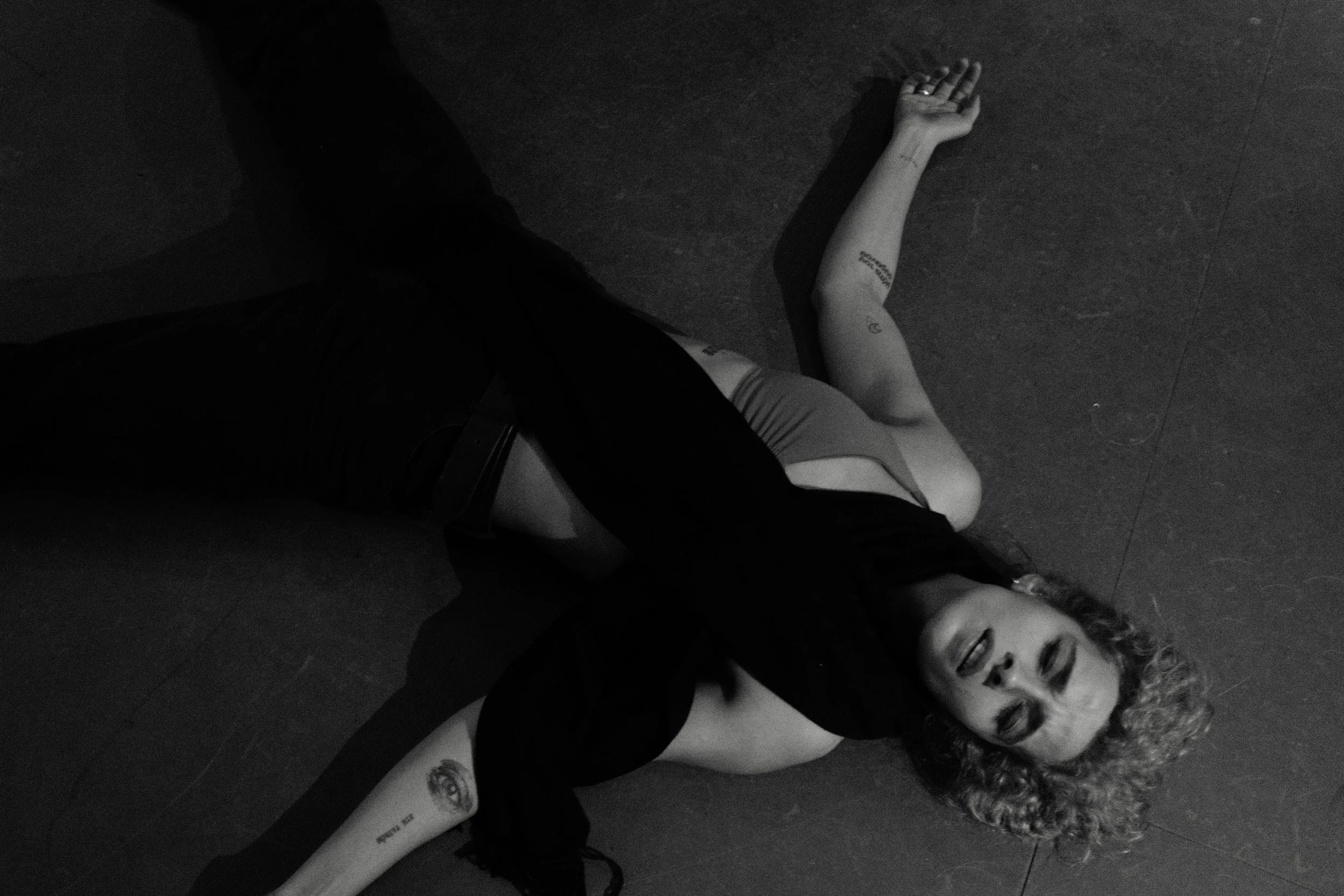 A woman with curly hair lying on the floor with a tattoo on her arm, smiling with her eyes closed. She is wearing a tank top and pants, and her head is tilted back.