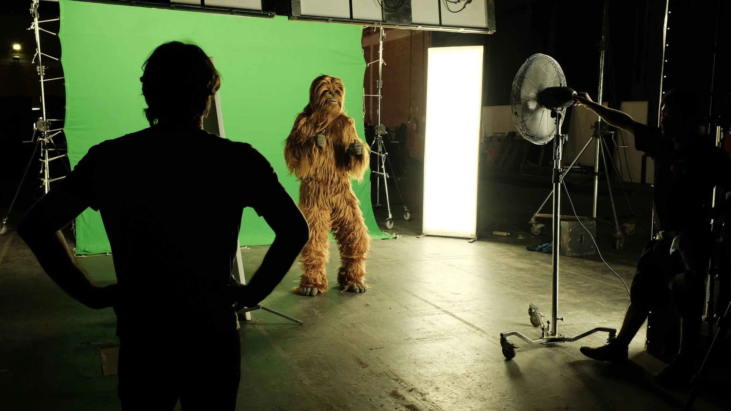 A behind-the-scenes view of a film or photo shoot with a person in a Chewbacca costume standing in front of a green screen, surrounded by studio equipment, with crew members working.