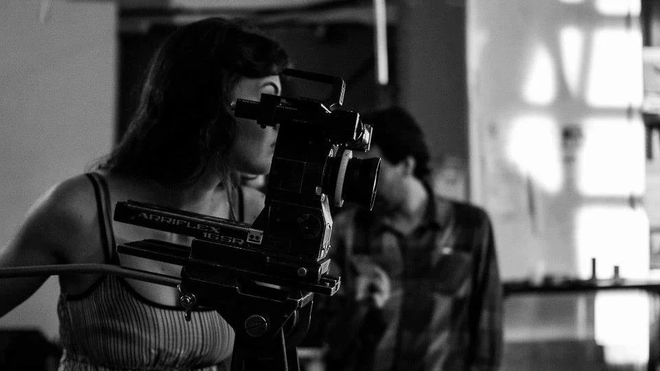 A woman operating a film camera in a studio or set, with another person in the background.