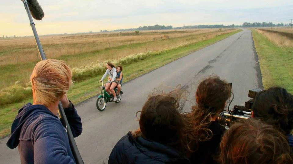 A behind-the-scenes shot on a film set showing a group of people watching a shot of two children riding a bicycle on a rural road, with a person holding a boom microphone and another person using a camera.