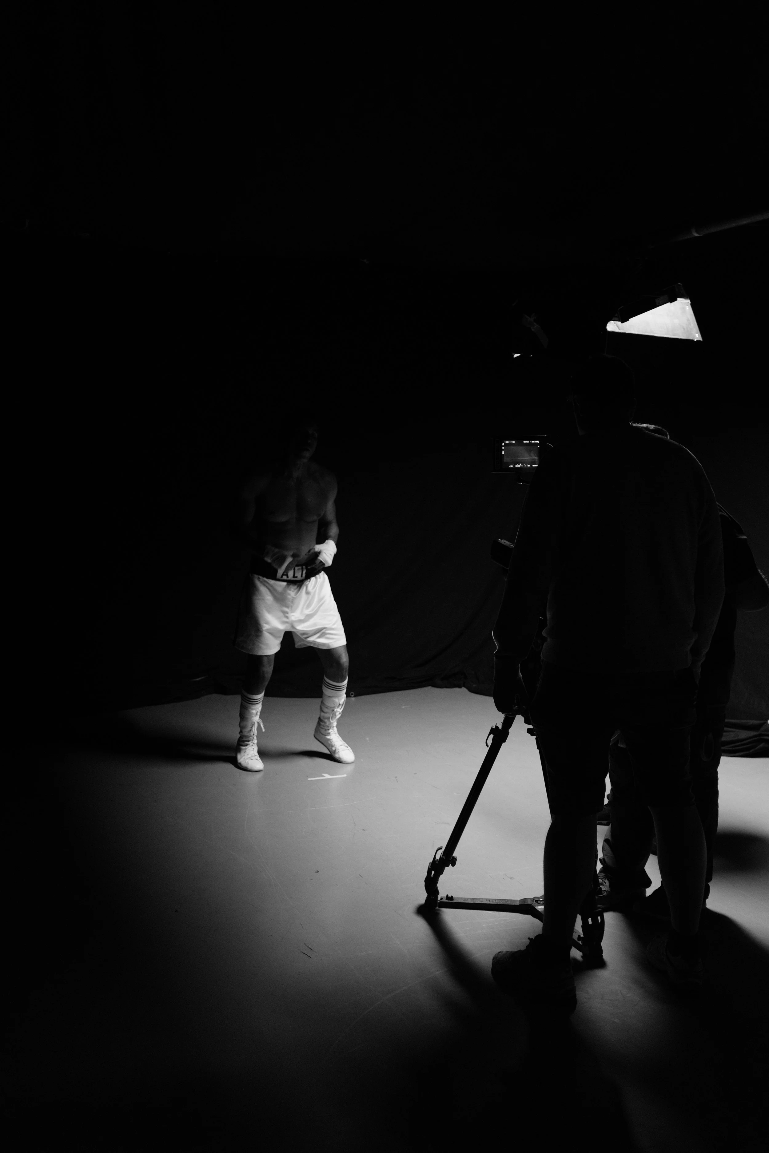Black and white image of a photo shoot in a studio. A shirtless male model dressed in white shorts, socks, and sneakers stands on a marked spot, while a photographer and assistant operate equipment nearby.