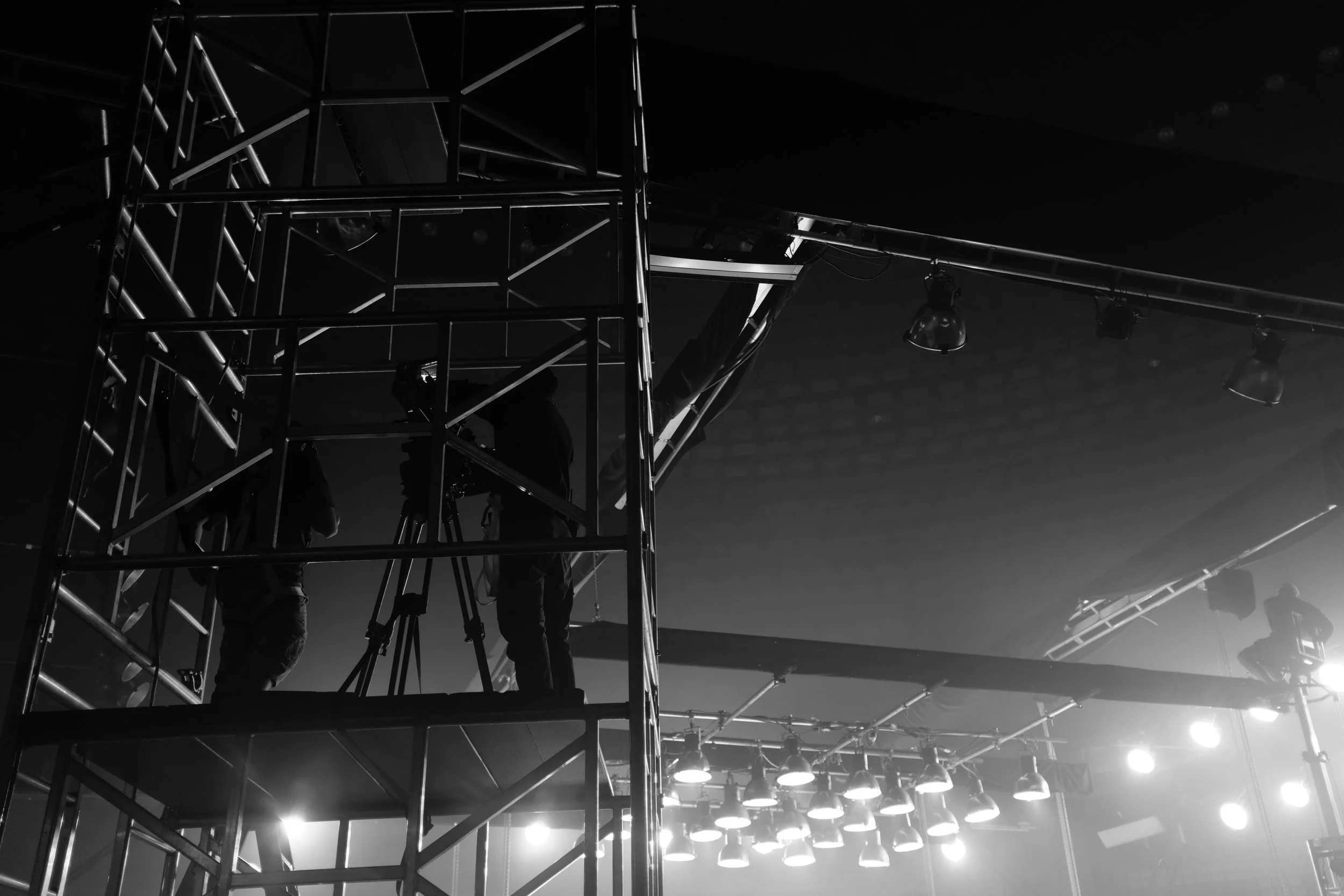 People working on a scaffolding structure in a dark indoor setting, with stage lights illuminating from below.