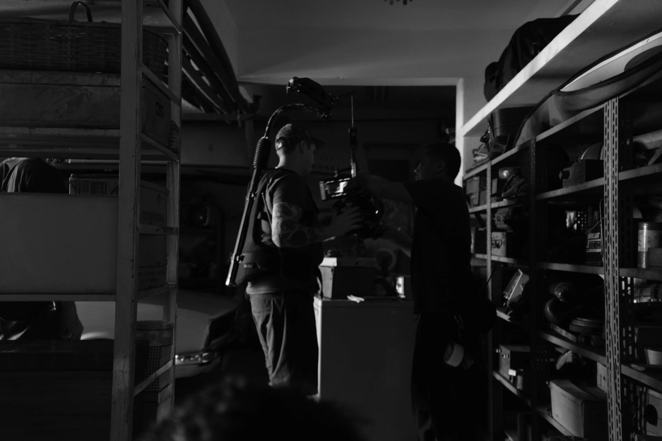 Two people are in a dimly lit storage room or warehouse, standing among shelves stocked with various items and equipment. One person appears to be holding or examining an object, while the other looks on. The setting suggests an interaction or discus