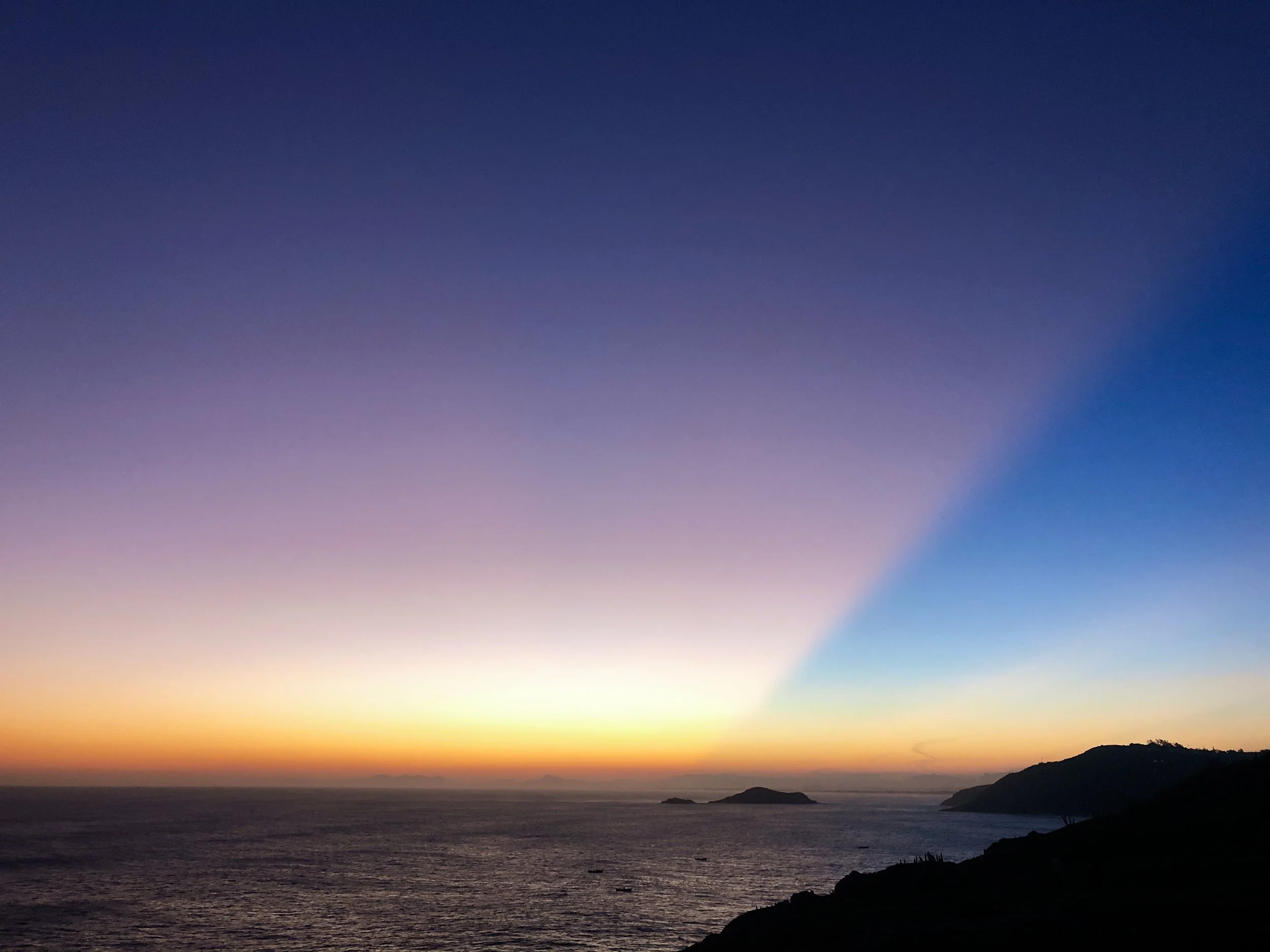 A sunset over the ocean with a colorful sky transitioning from orange and yellow near the horizon to purple and blue higher up. There are small islands or landmasses visible in the distance, and silhouette of a hillside or shoreline in the foreground