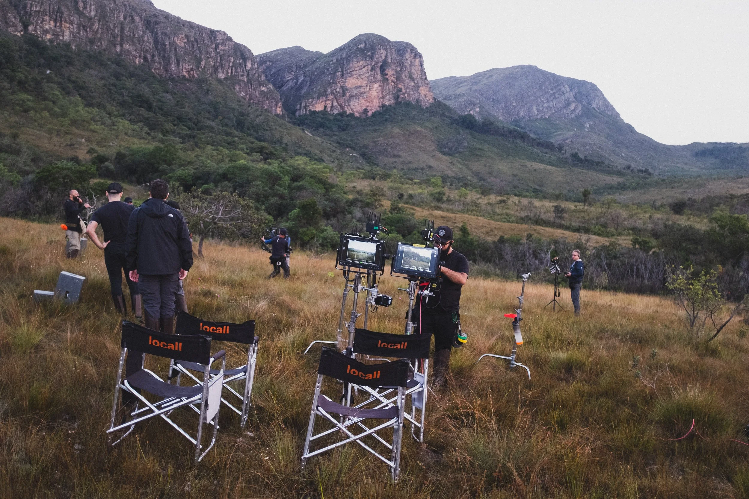 Group of people in an outdoor field with equipment on tripods, surrounded by grassy landscape and mountains in the background, used for filming or photography.