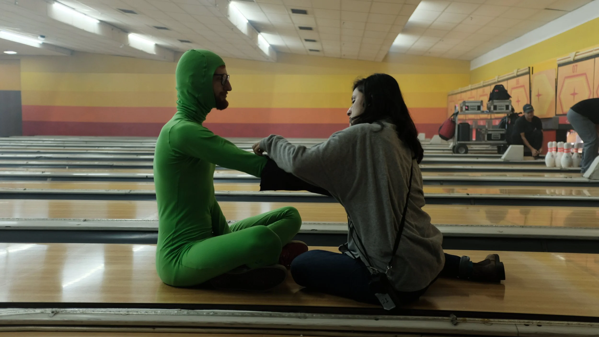 A person dressed in a green superhero costume with a hood and glasses sitting cross-legged at a bowling alley, holding hands with a woman in a gray hoodie who is kneeling and smiling.