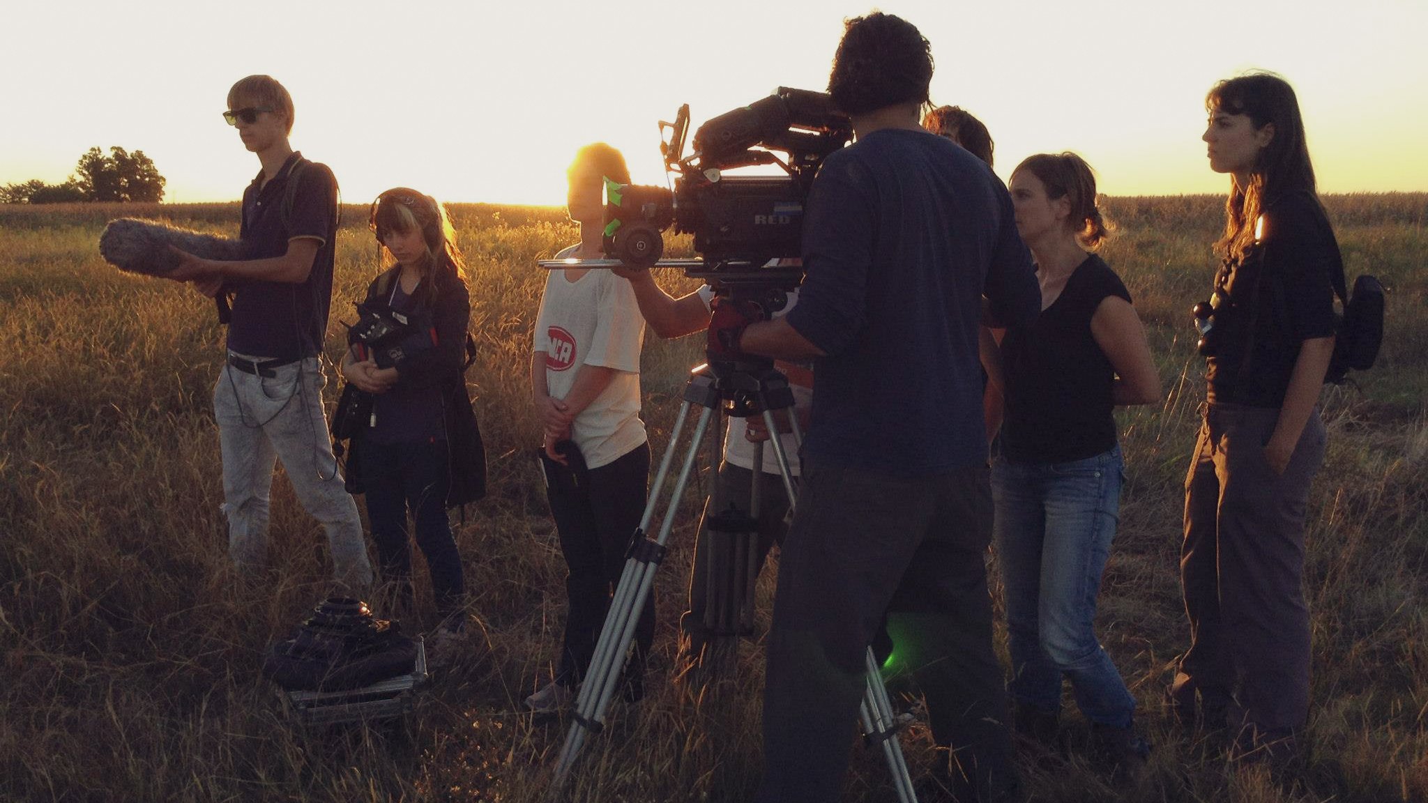 A film crew working outdoors during sunset, with a camera on a tripod, several people, and a grassy field.