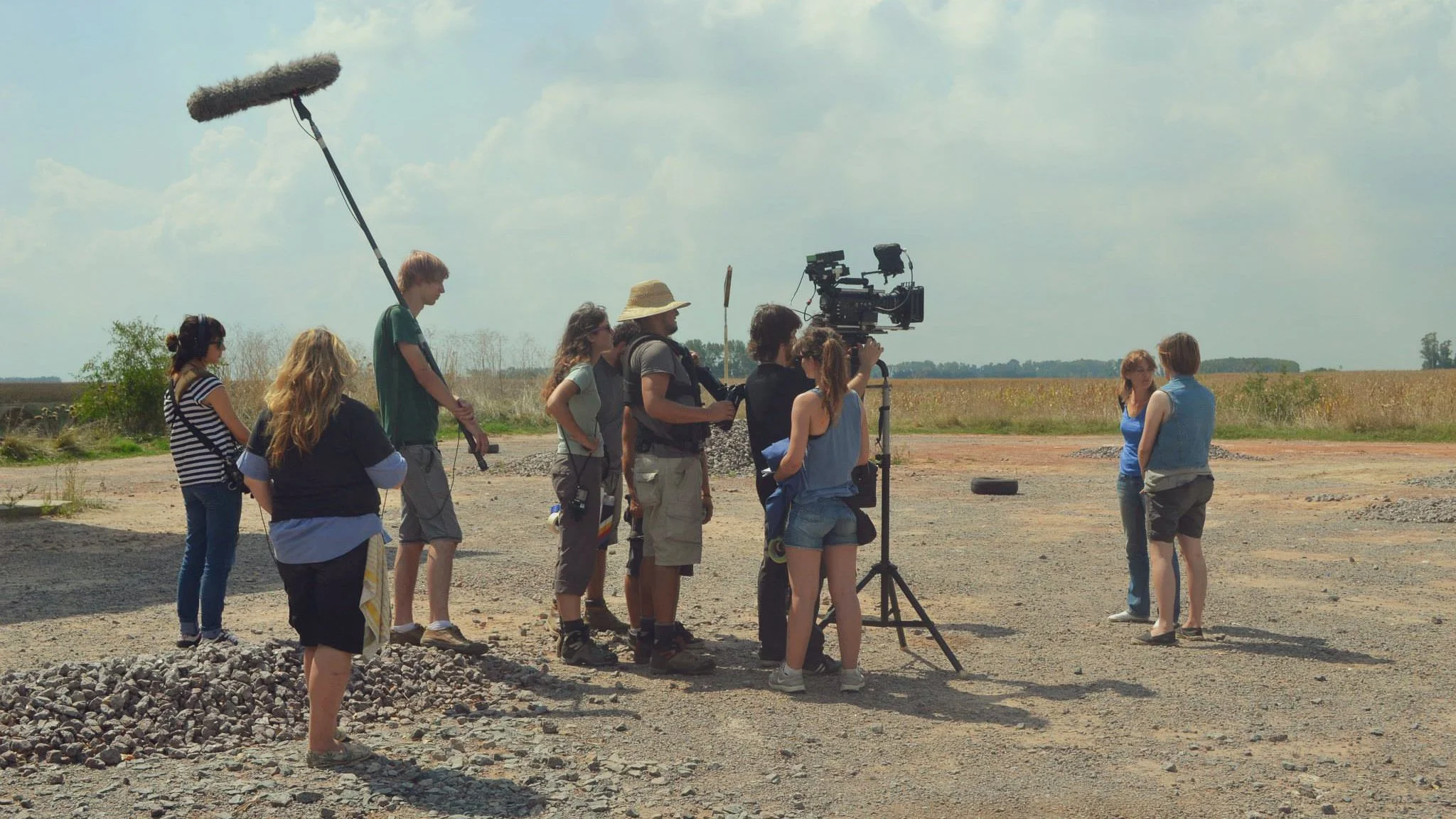 A film crew filming a scene outdoors on a dirt area with a wide-open field and cloudy sky in the background. The crew includes people operating a camera and sound equipment, with two women talking to each other on the right side of the image.