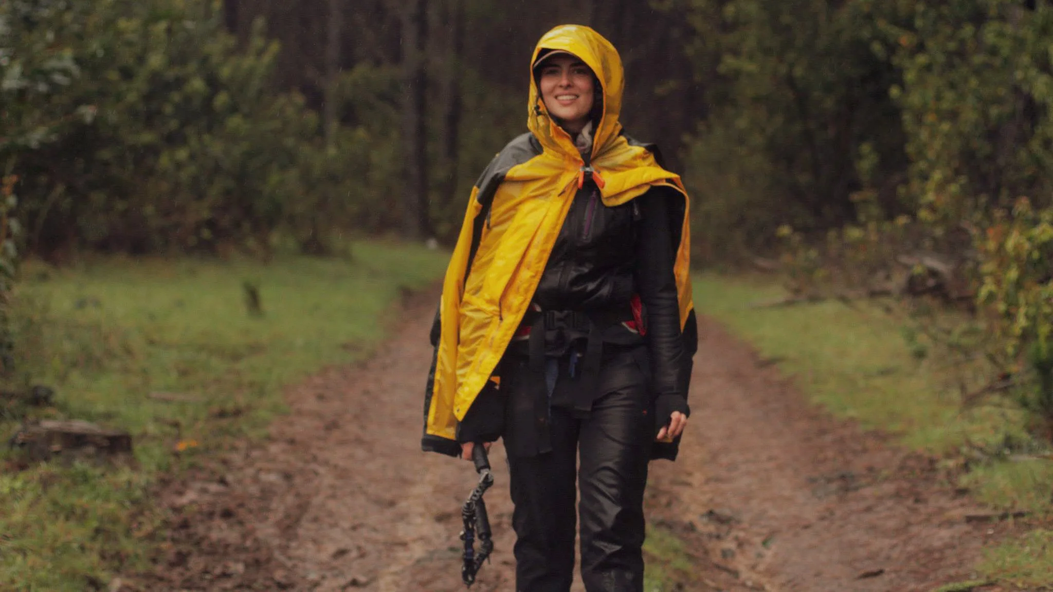 A woman dressed in outdoor gear with a yellow raincoat and black pants walking on a dirt trail in a forested area, smiling with her hands at her sides.