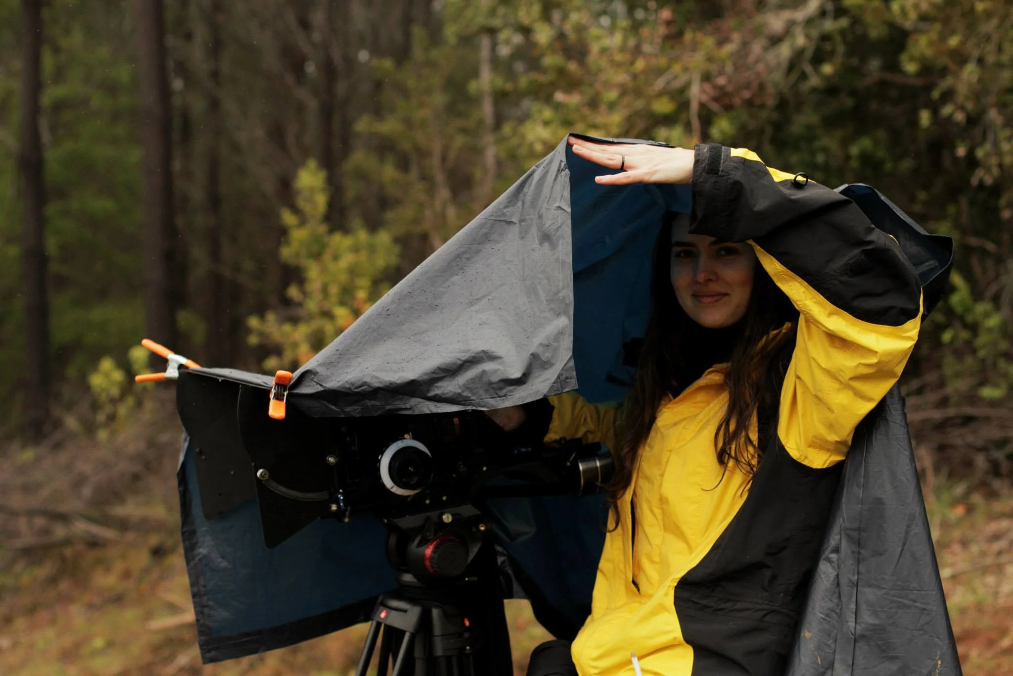 A woman dressed in a yellow and black rain jacket adjusting her hat, standing outdoors in front of a forested background, near a mounted camera on a tripod.