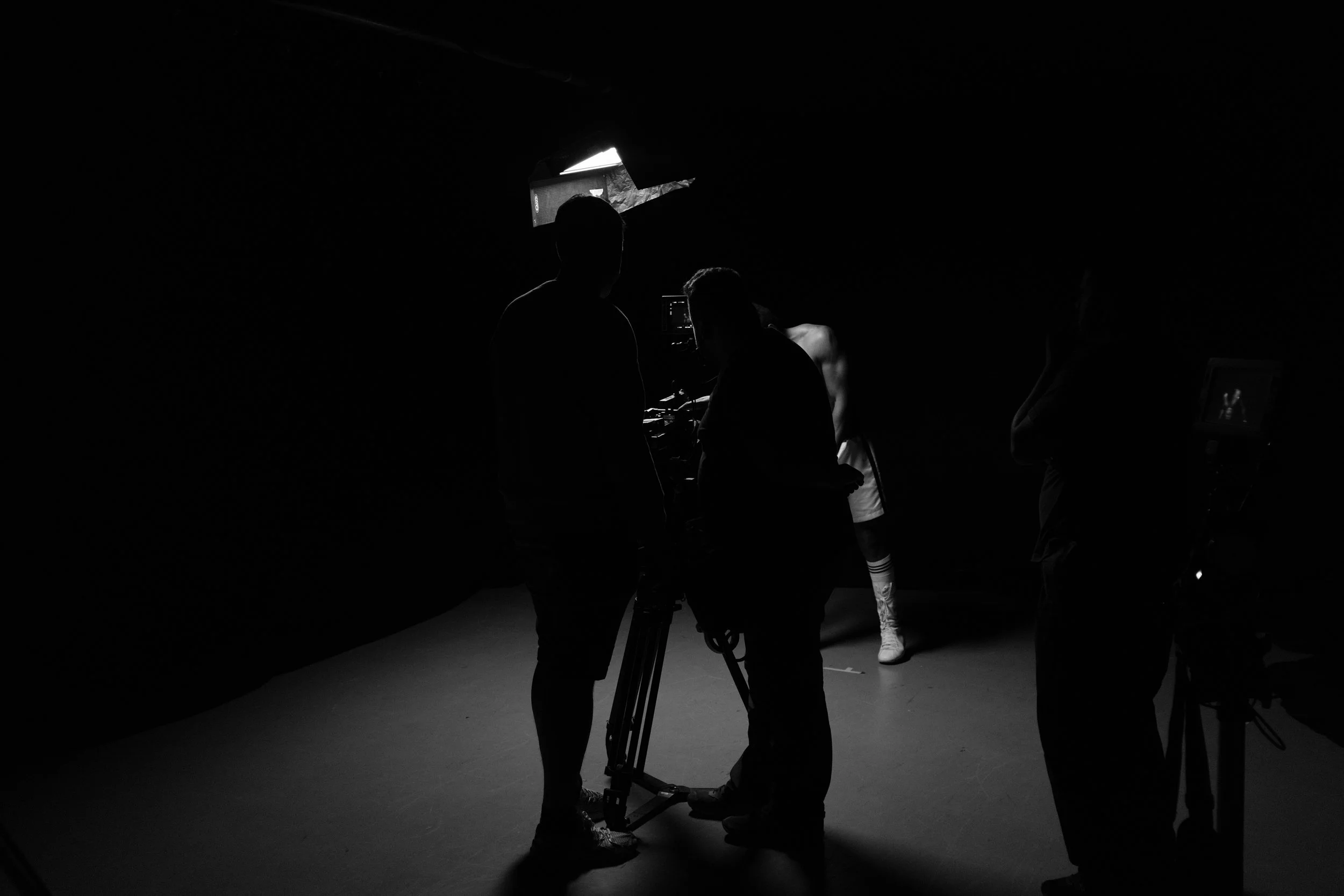 Silhouettes of people behind the scenes of a video shoot in a dark studio, with a person adjusting equipment and a model in the background.