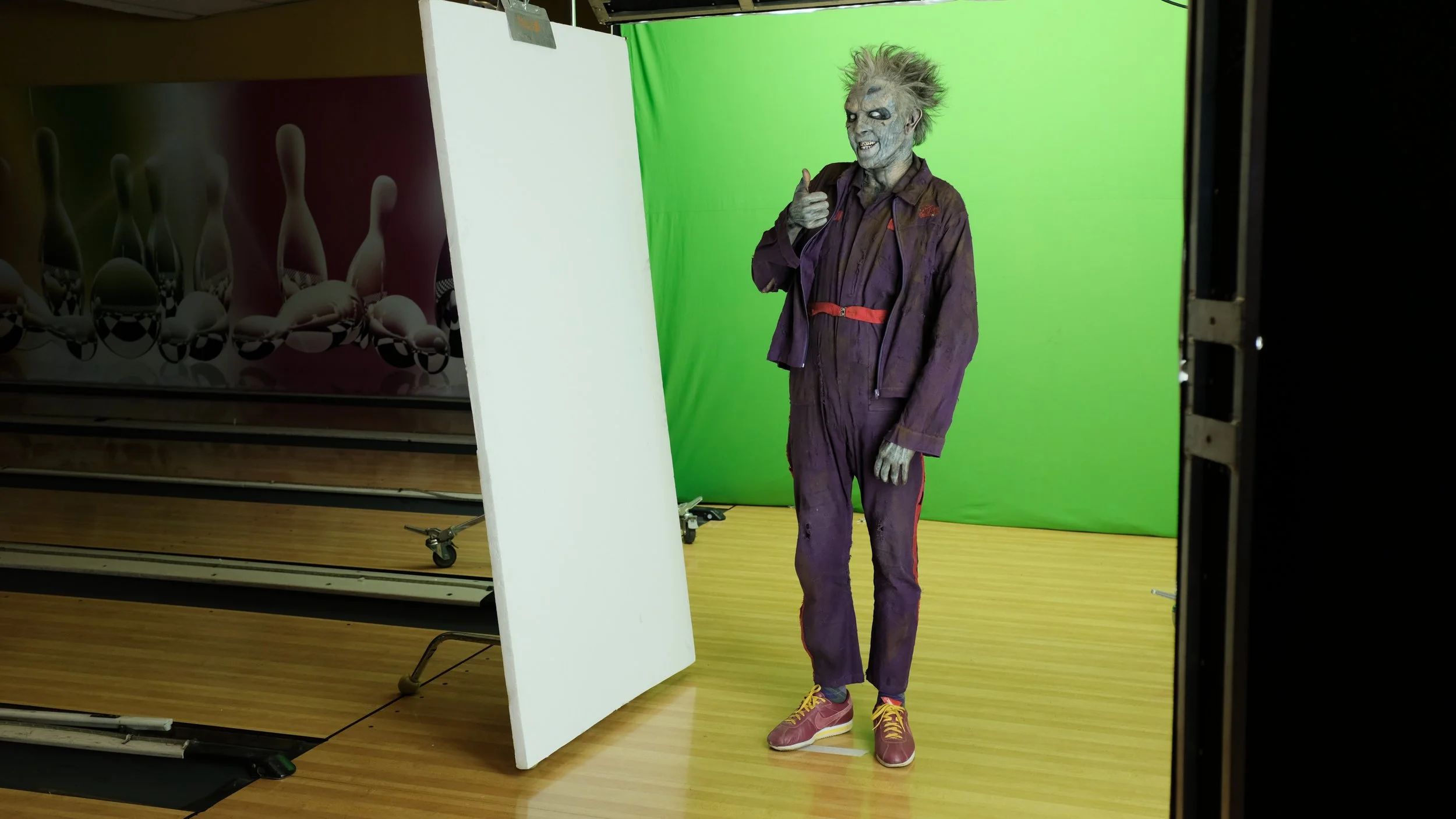 A person dressed as a zombie wearing a purple jumpsuit with red accents and yellow sneakers giving a thumbs-up in front of a green screen at a photo studio.