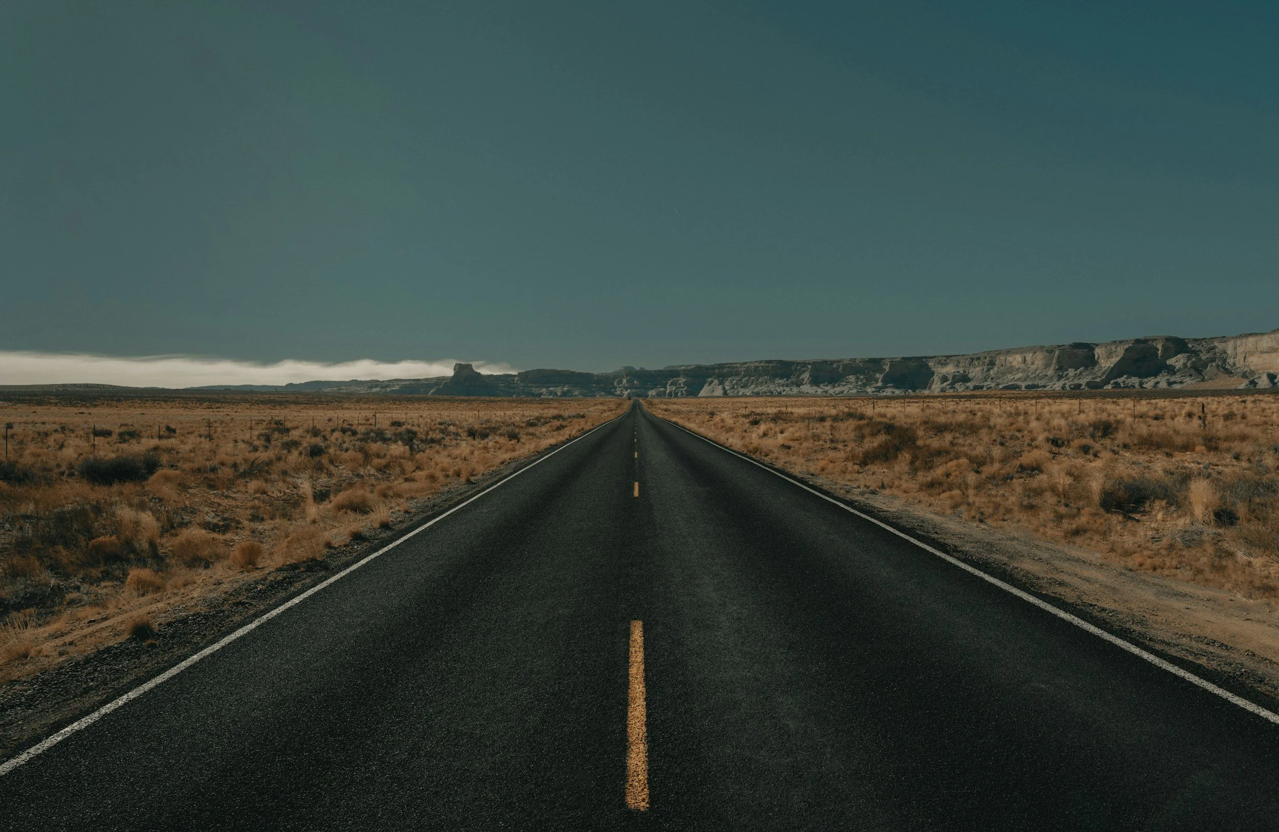 A straight two-lane highway running through a desert landscape with mountains in the distance under a cloudy sky.