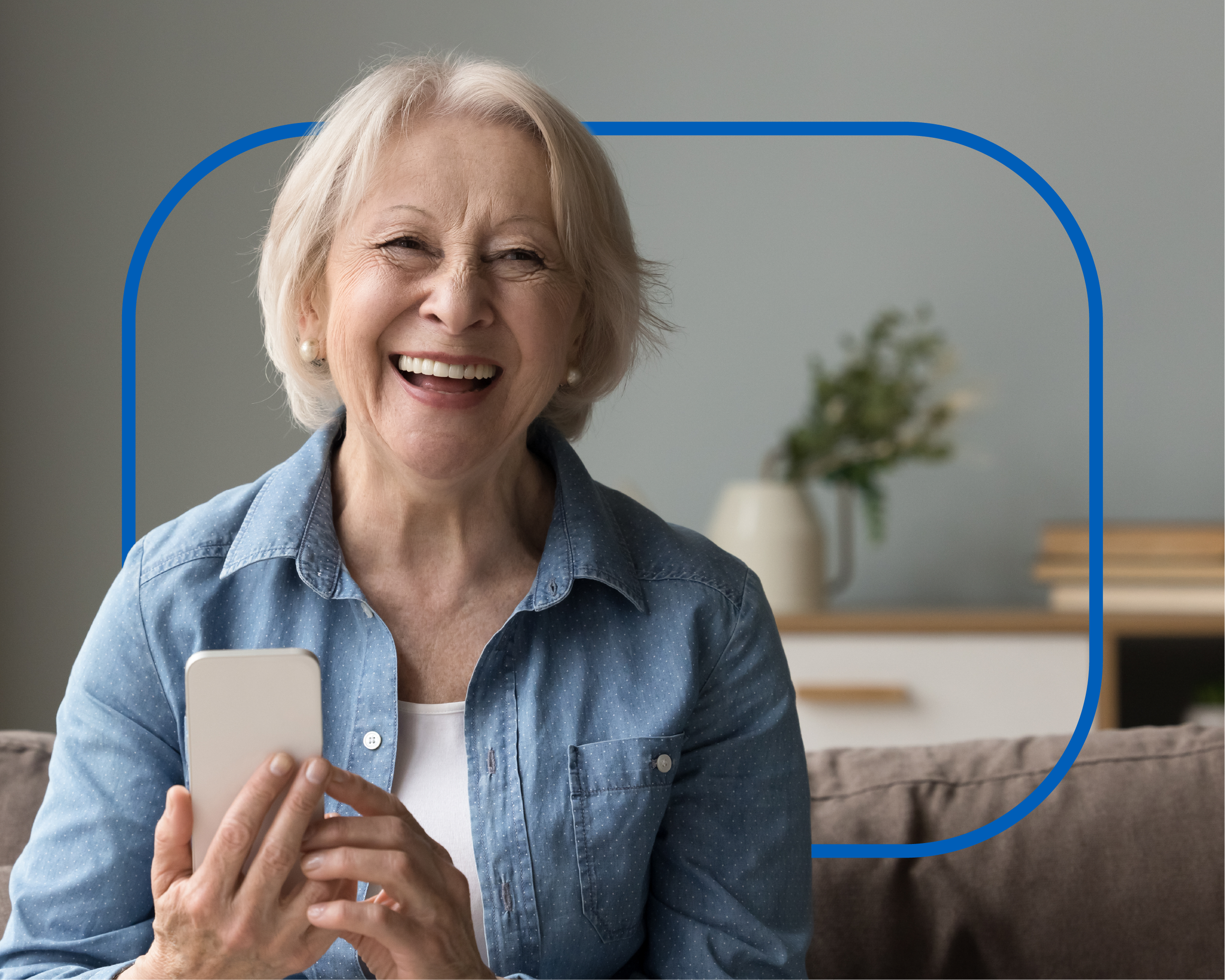 An older woman with short white hair, wearing a blue denim shirt and pearl earrings, smiling and looking at her phone while sitting in a cozy room.