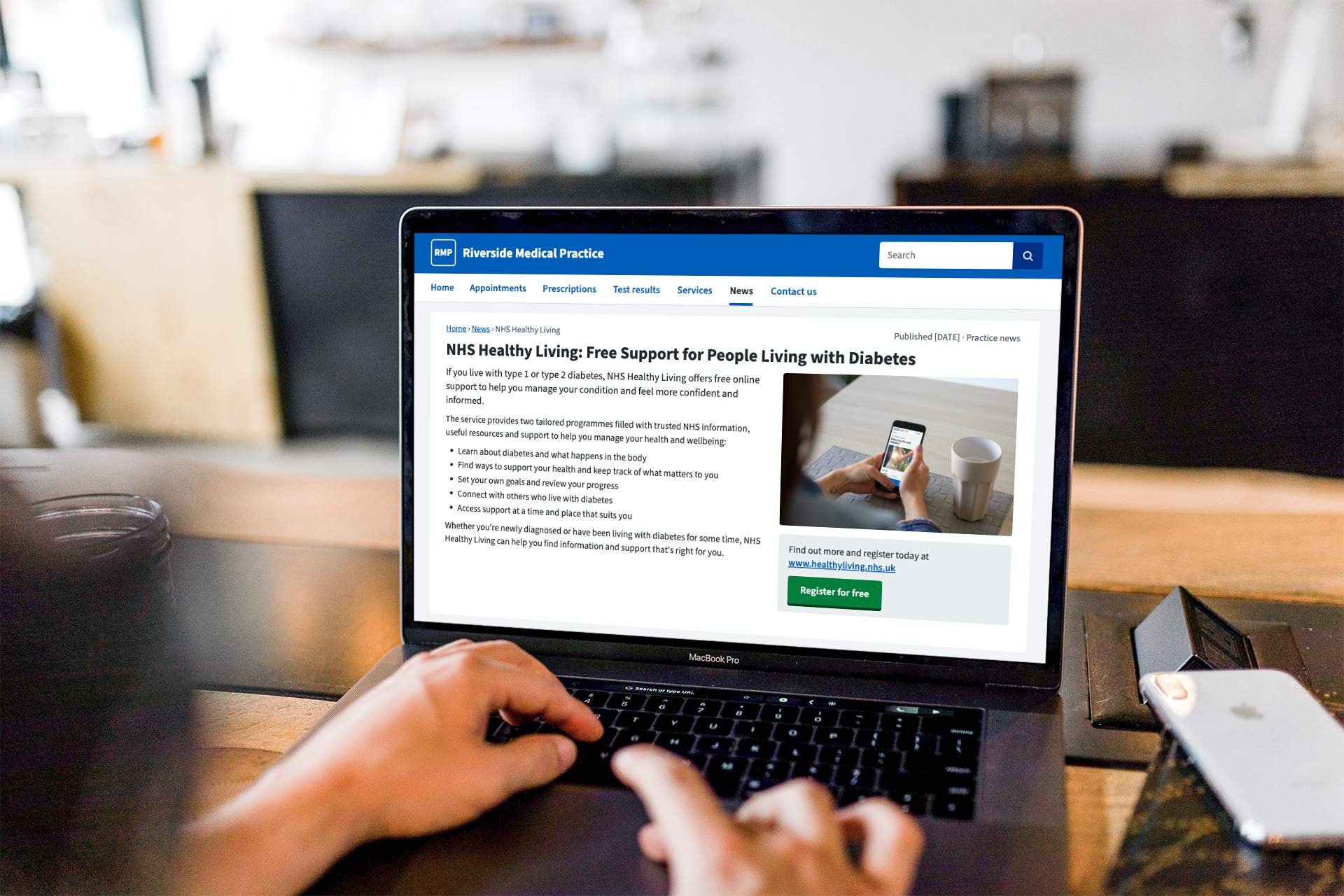 A person using a MacBook Pro laptop at a brown wooden table in a well-lit room with a blurred background. The laptop screen shows a health website about NHS Healthy Living support for people with diabetes.