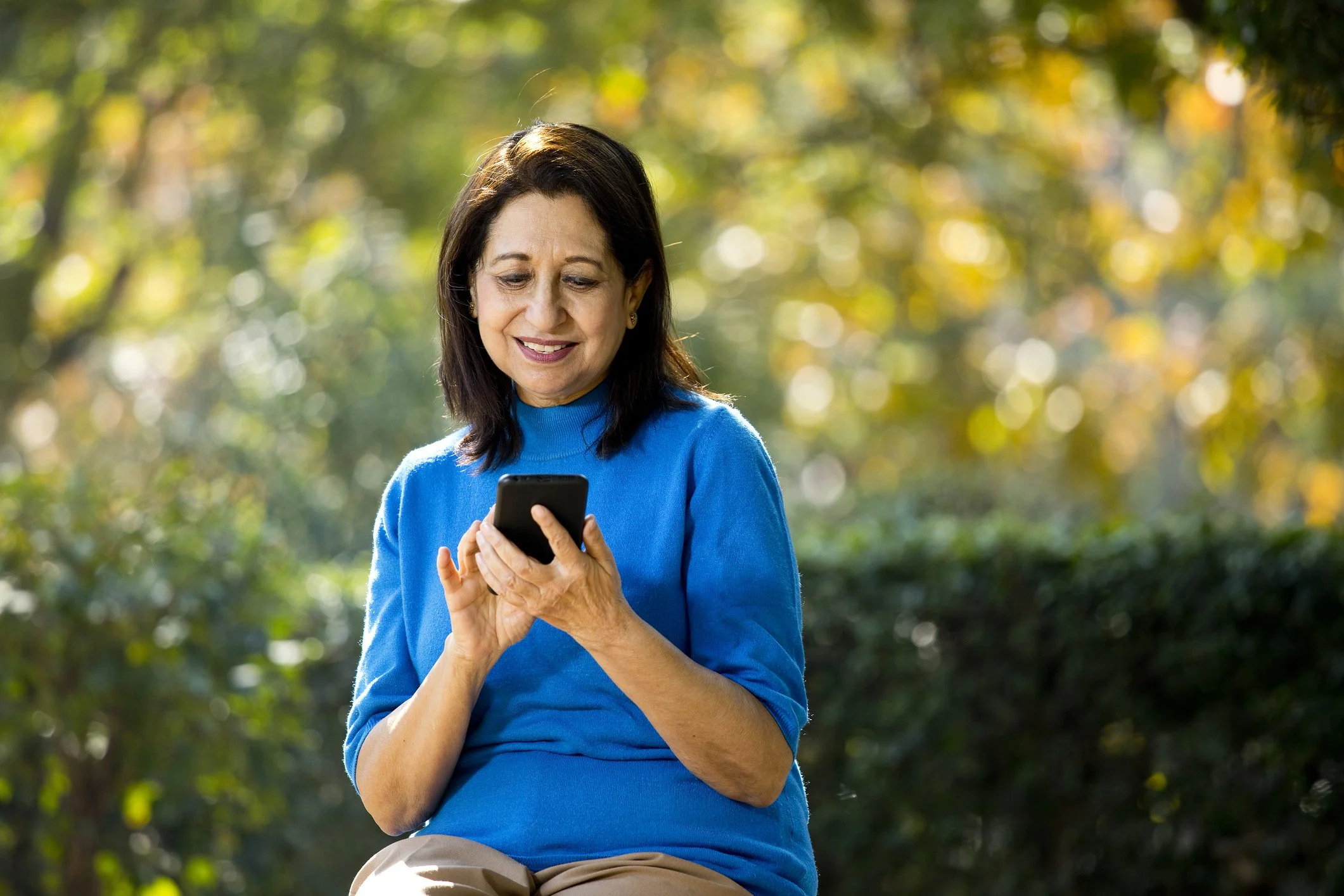 A woman in a blue sweater sitting outdoors looking at her phone in a park with green and yellow foliage.