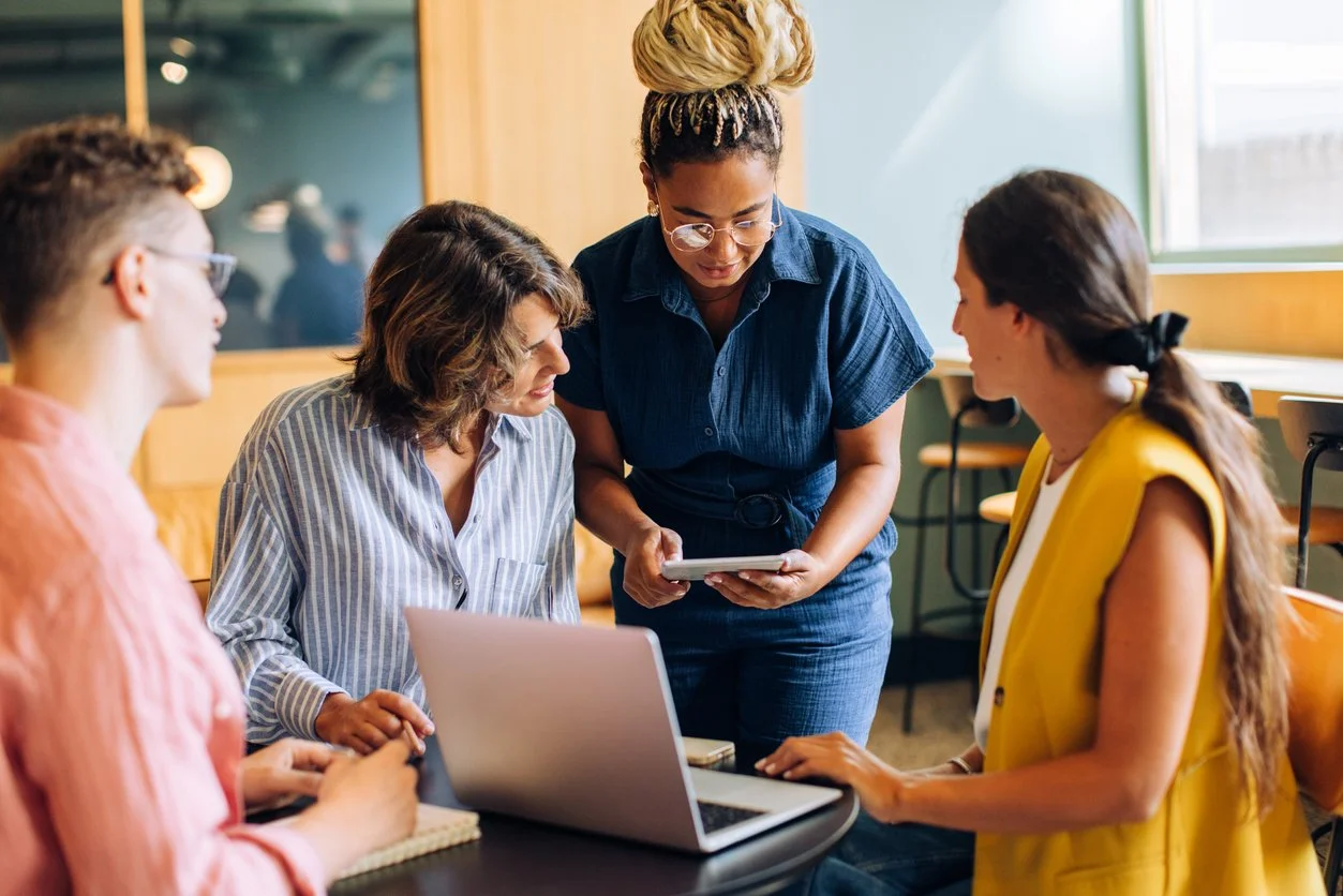 Four women gathered around a laptop in a meeting, one woman standing and looking at her phone while the others are seated and looking at her.