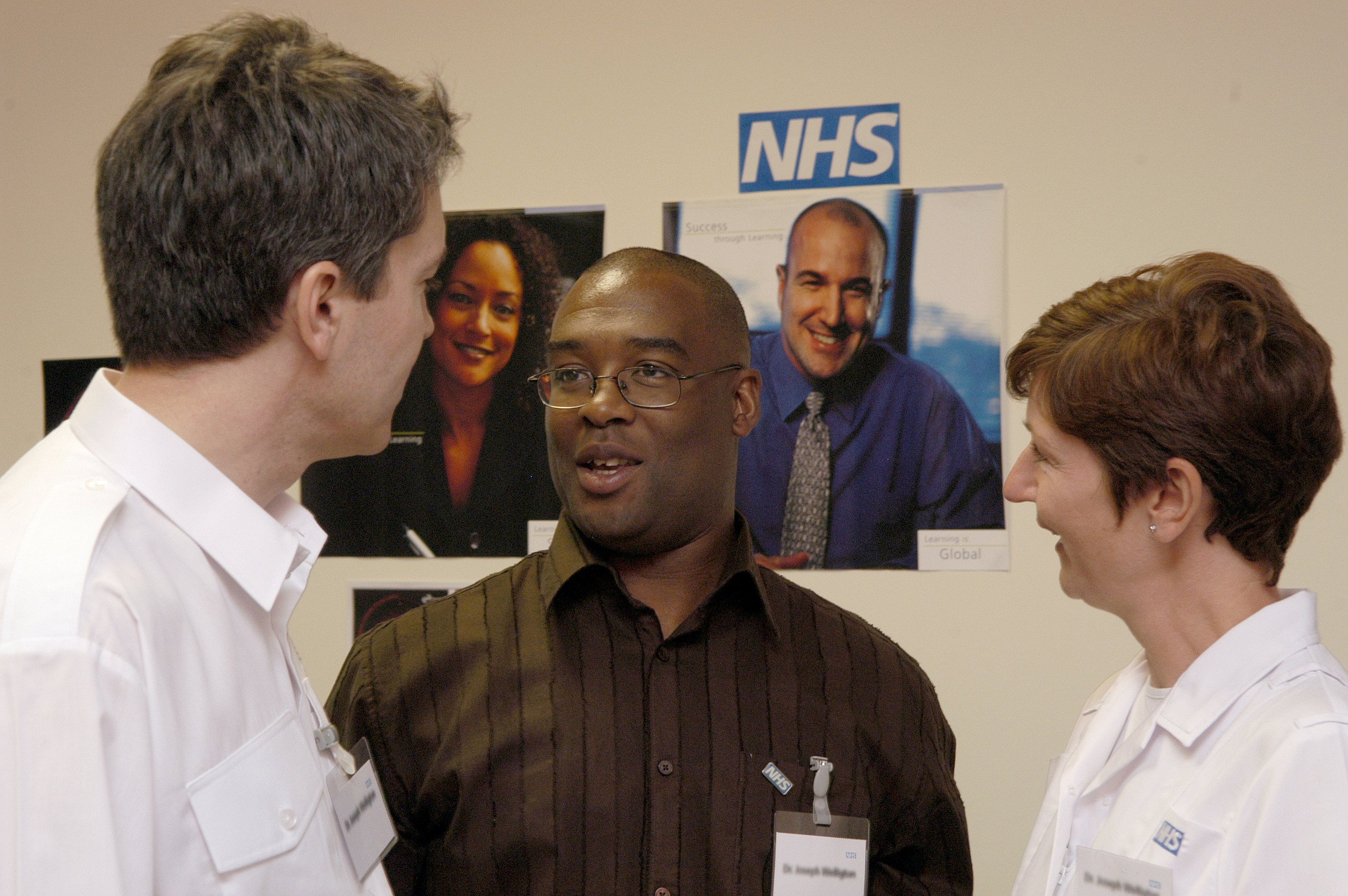 Three people having a conversation in front of a wall with NHS posters. The man in the middle is wearing glasses and a brown striped shirt, talking to two women in white uniforms.