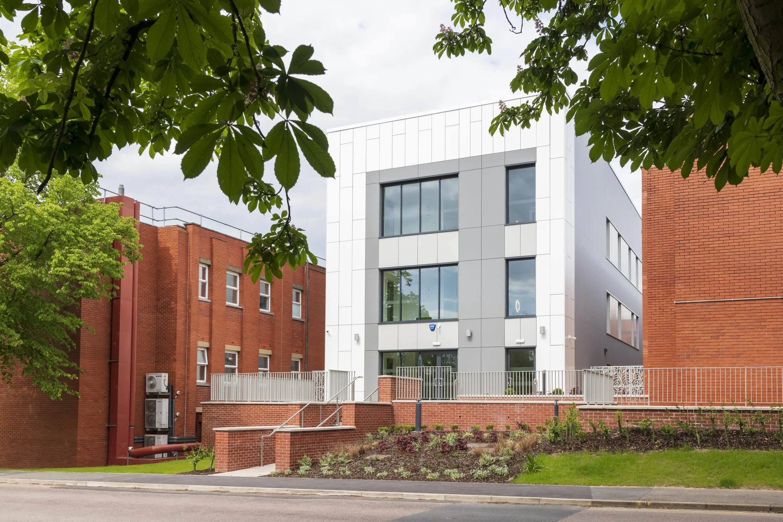 Modern white building with large windows, surrounded by red brick and greenery with trees and garden, under a cloudy sky.