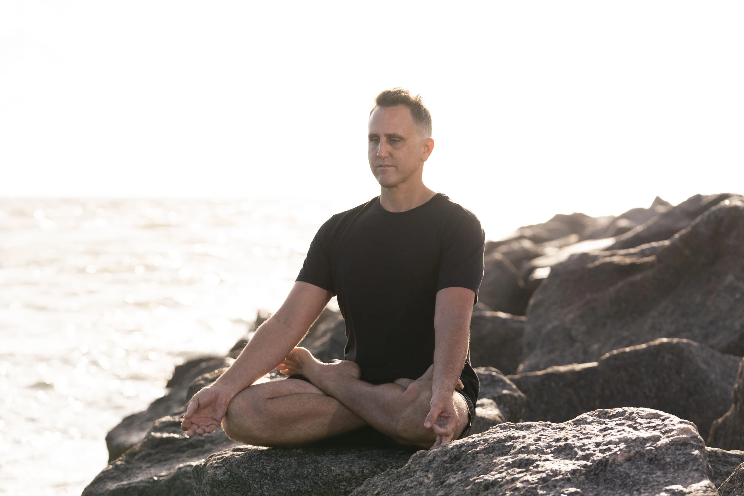 A man sitting in lotus pose and arms down by his side wearing shorts and.a black t-shirt sitting on rocks with a whiter sky.