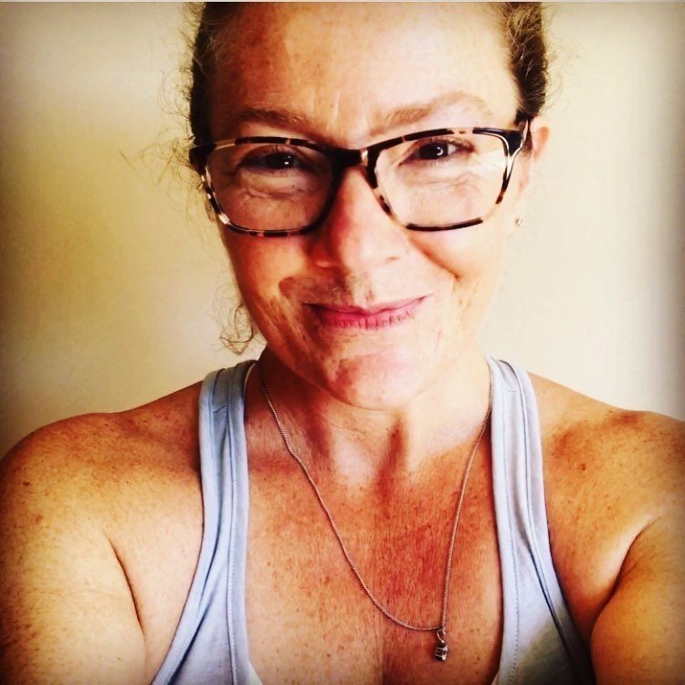 a selfie of a women smiling at the camera with a singlet top on upper arm, shoulder part of chest and collar bones showing and most  of head  wearing black glasses and a necklace smiling. fair skin with lots of freckles and moles.