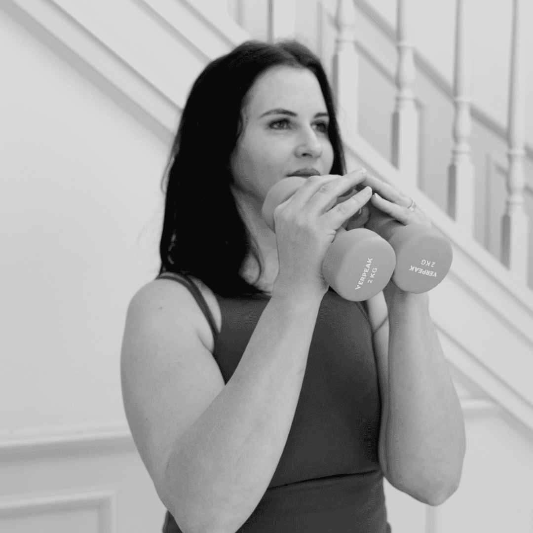 A woman with dark hair, wearing a sleeveless top, holding two dumbbells with 'VERPEAK 2 KG' written on them, in front of a staircase with white spindles.