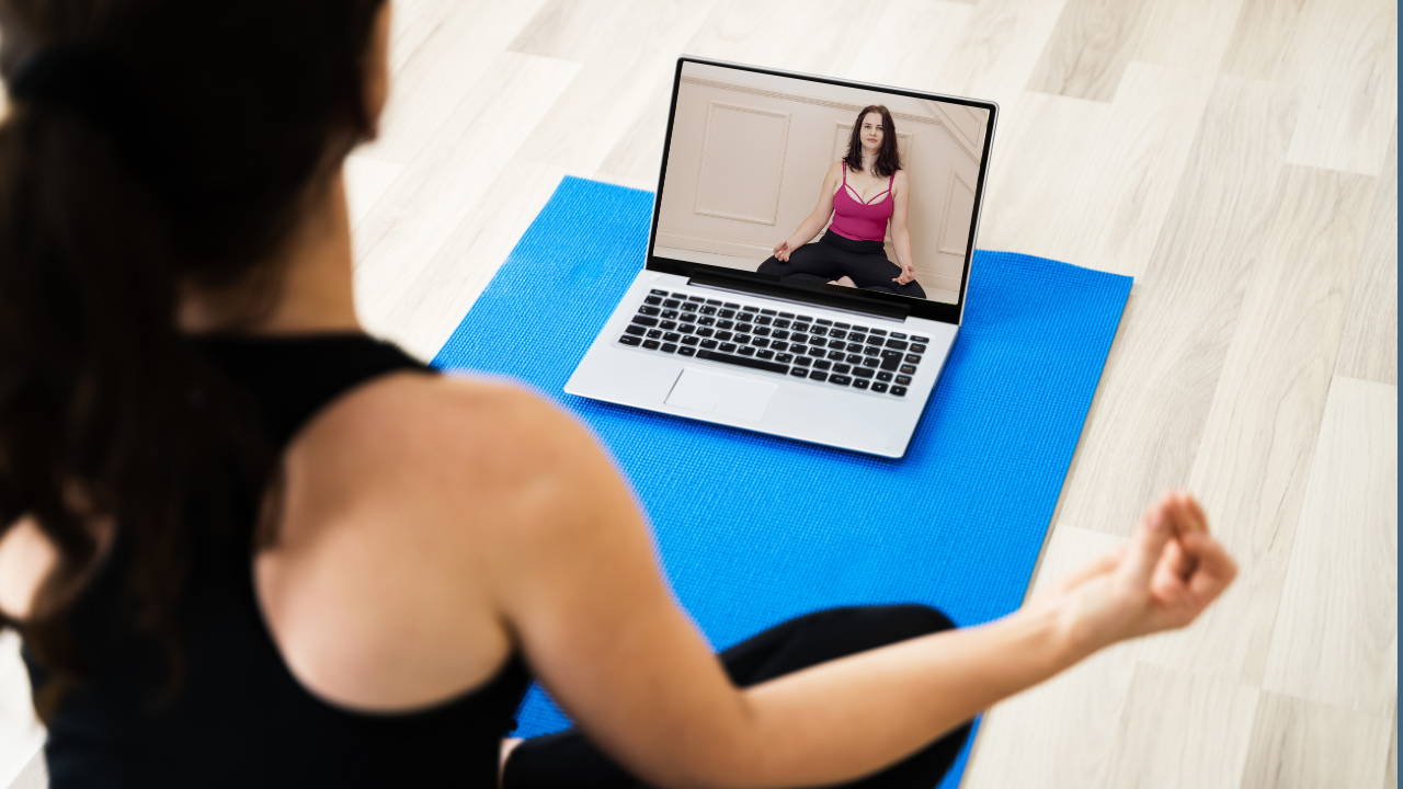 A woman practicing yoga on a blue mat while participating in a virtual yoga class via laptop in a bright indoor setting.
