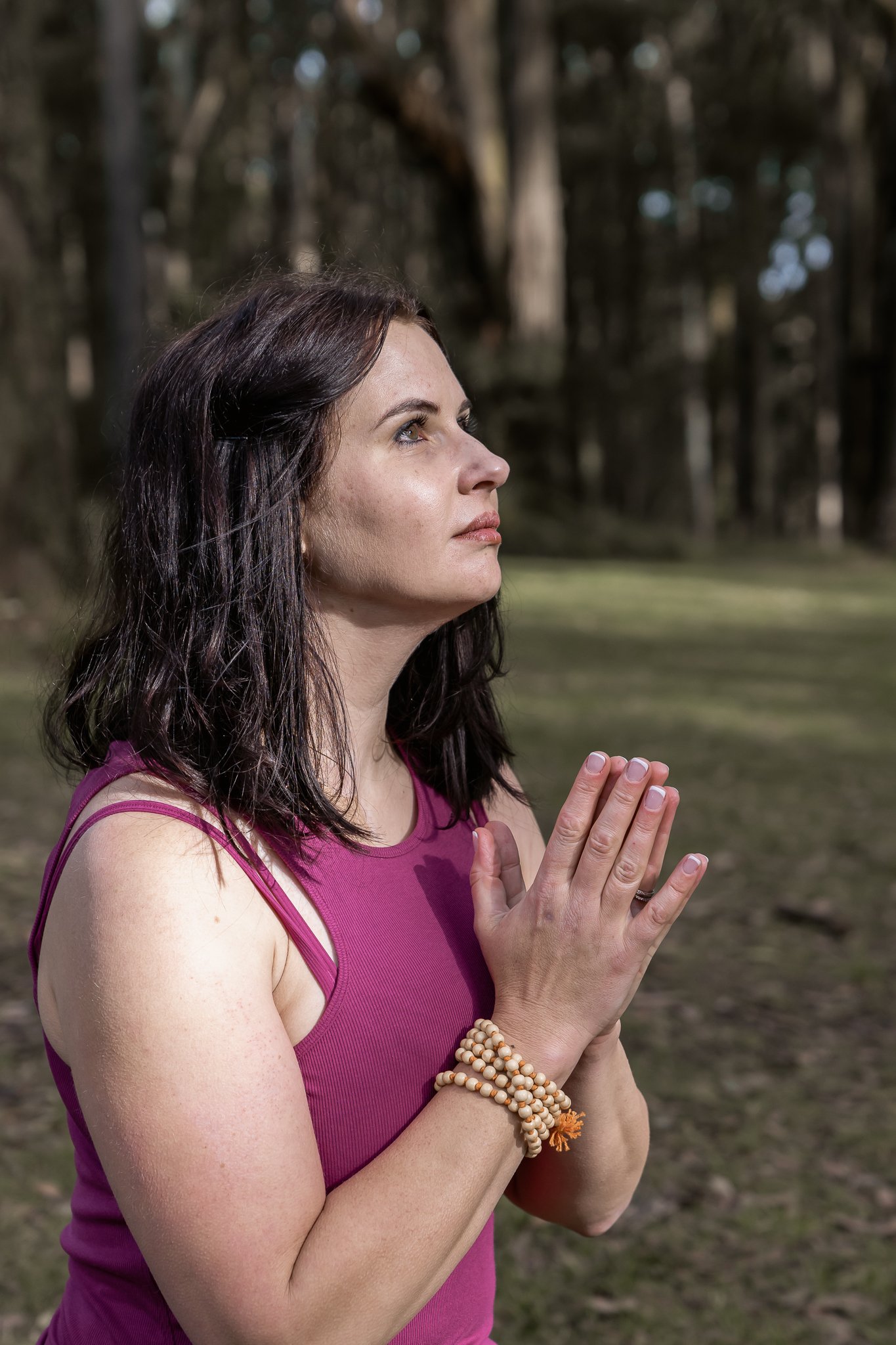 woman with dark shoulder length hair wearing a pink yoga top and jape meditation beads with hands in prayer position side on looking softly above with grass and trees in the background